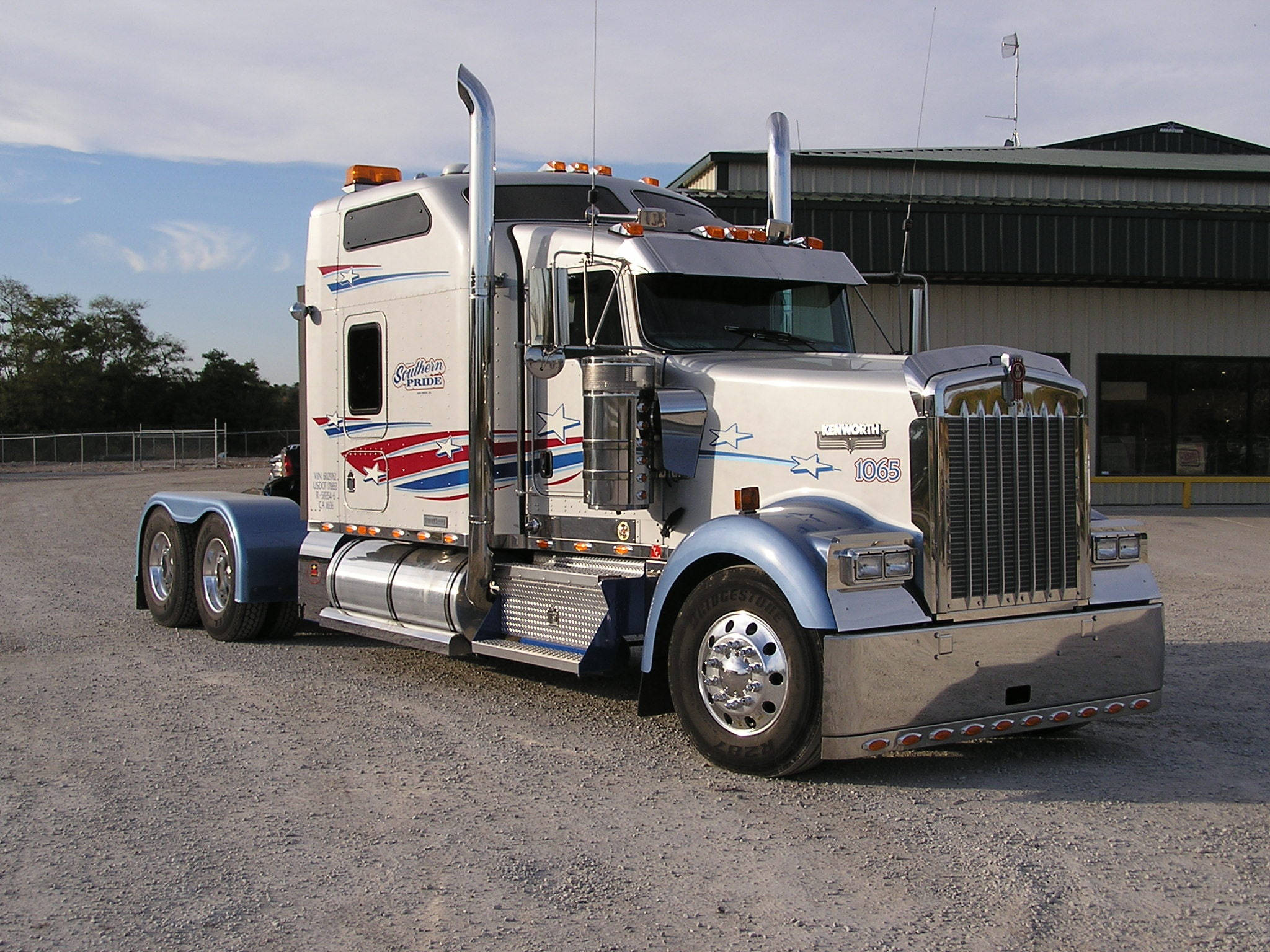 Kenworth W900 桌布 壁紙 — 一輛閃亮的銀色 Kenworth 半掛卡車，帶有愛國紅白藍貼花，停在倉庫附近的礫石上。