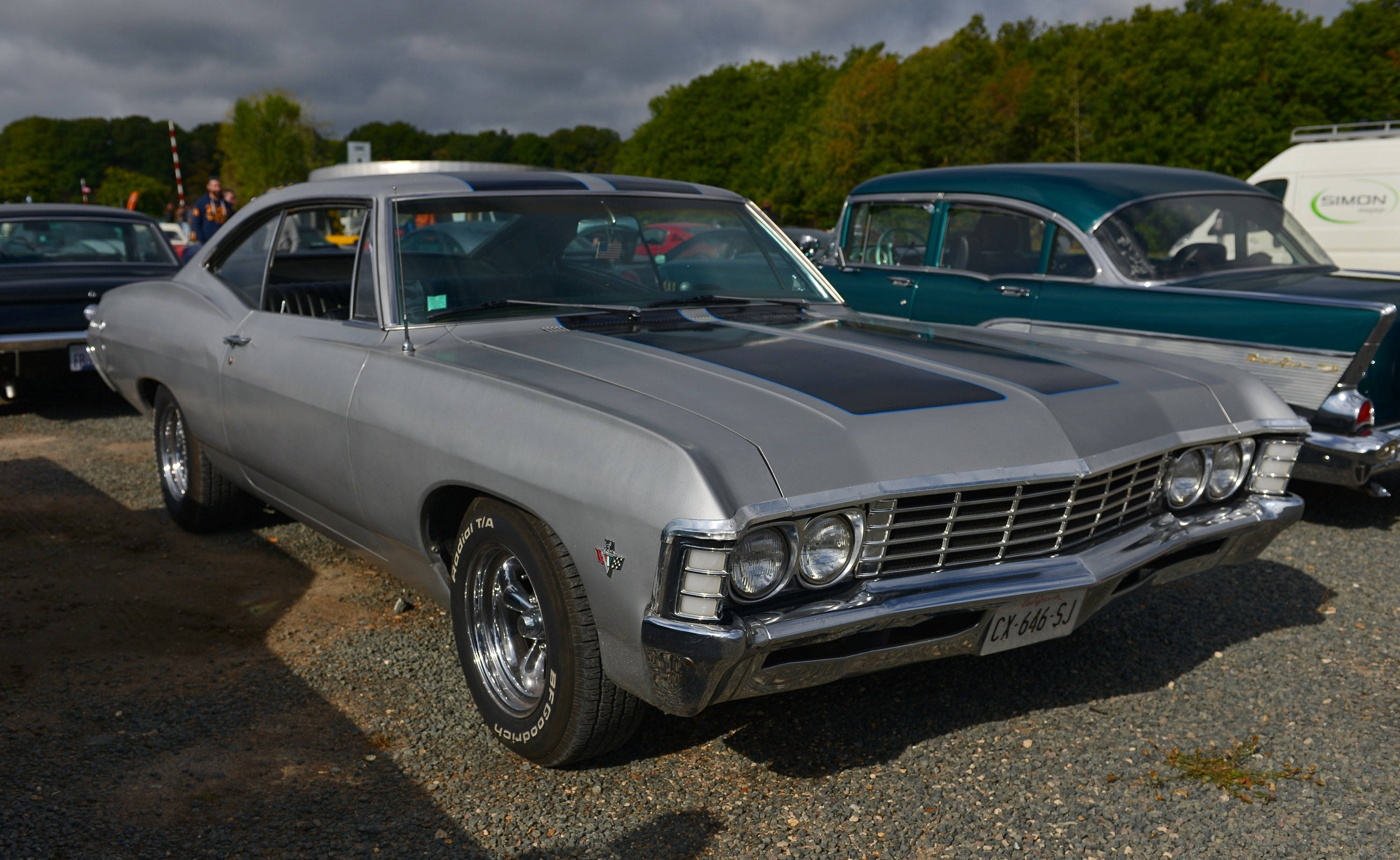 chevrolet impala 1967 桌布 壁紙 — 一輛閃亮的銀色經典肌肉車，帶有黑色引擎蓋條紋，停在碎石地上，在陰天下的車展中。