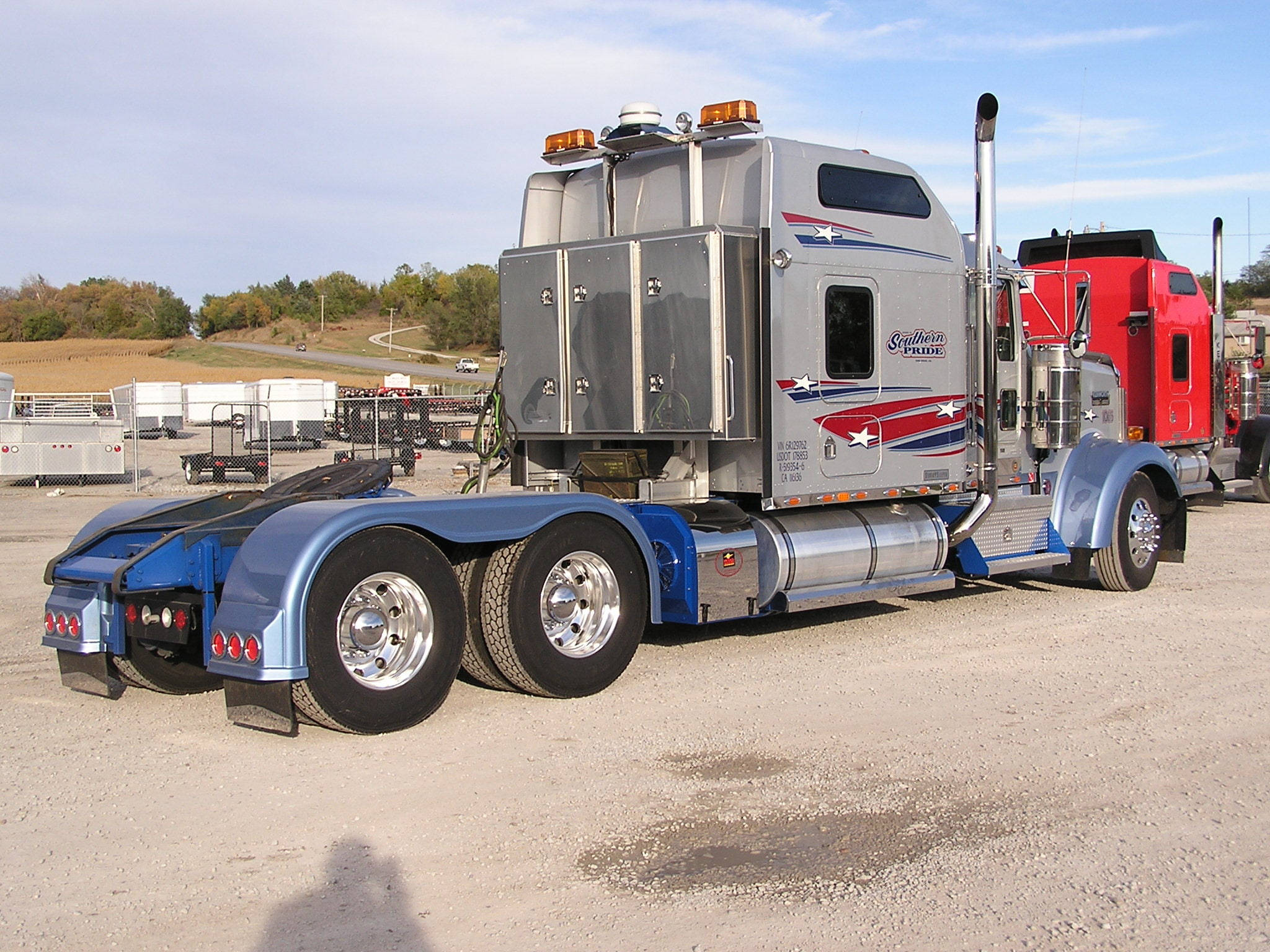 Kenworth W900 桌布 壁紙 — 一輛閃亮的銀藍色半掛卡車帶有紅白藍愛國貼紙，停在陽光明媚的戶外碎石場上。