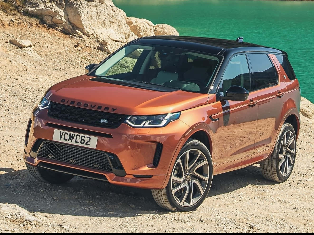 Land Rover Discovery 桌布 壁紙 — 一輛閃亮的銅色 Land Rover Discovery 停放在岩石地形上，旁邊是充滿活力的綠松石色湖泊，在明亮的日光下。