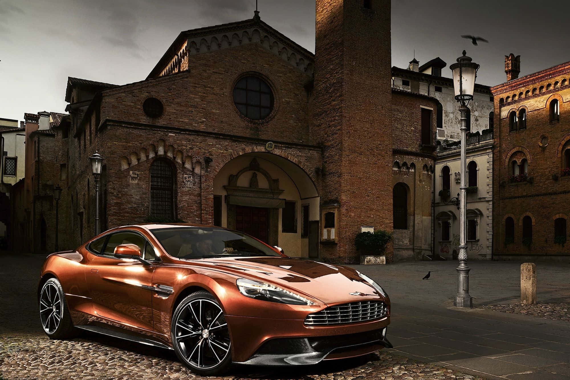 Aston Martin Rapide S 桌布 壁紙 — 一輛閃亮的銅色Aston Martin跑車停泊在鵝卵石廣場上，前方是歷史悠久的磚砌建築，天空陰沉多雲。