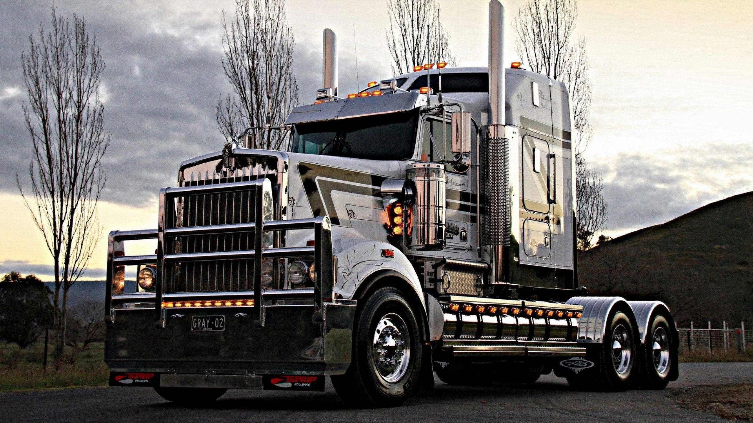 Kenworth 桌布 壁紙 — 一輛閃亮的鍍鉻半掛卡車配備雙排氣管，在黃昏時分停放在鄉村道路上，兩旁是光禿禿的樹木，天空呈現柔和的漸變色。