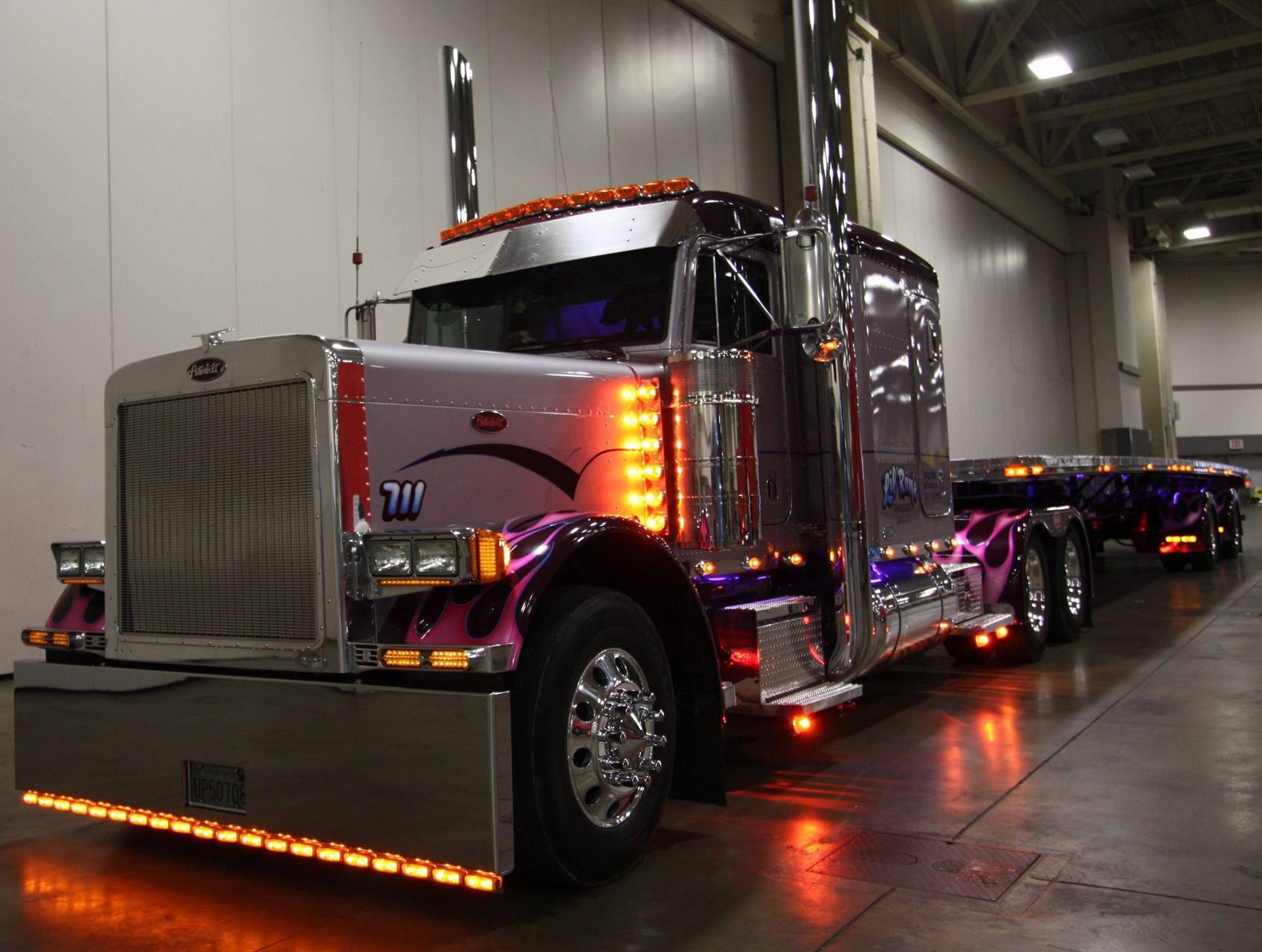 Peterbilt 桌布 壁紙 — 一輛閃亮的鍍鉻和紫色定制半掛卡車，帶有發光的橙色燈光，停在倉庫中。