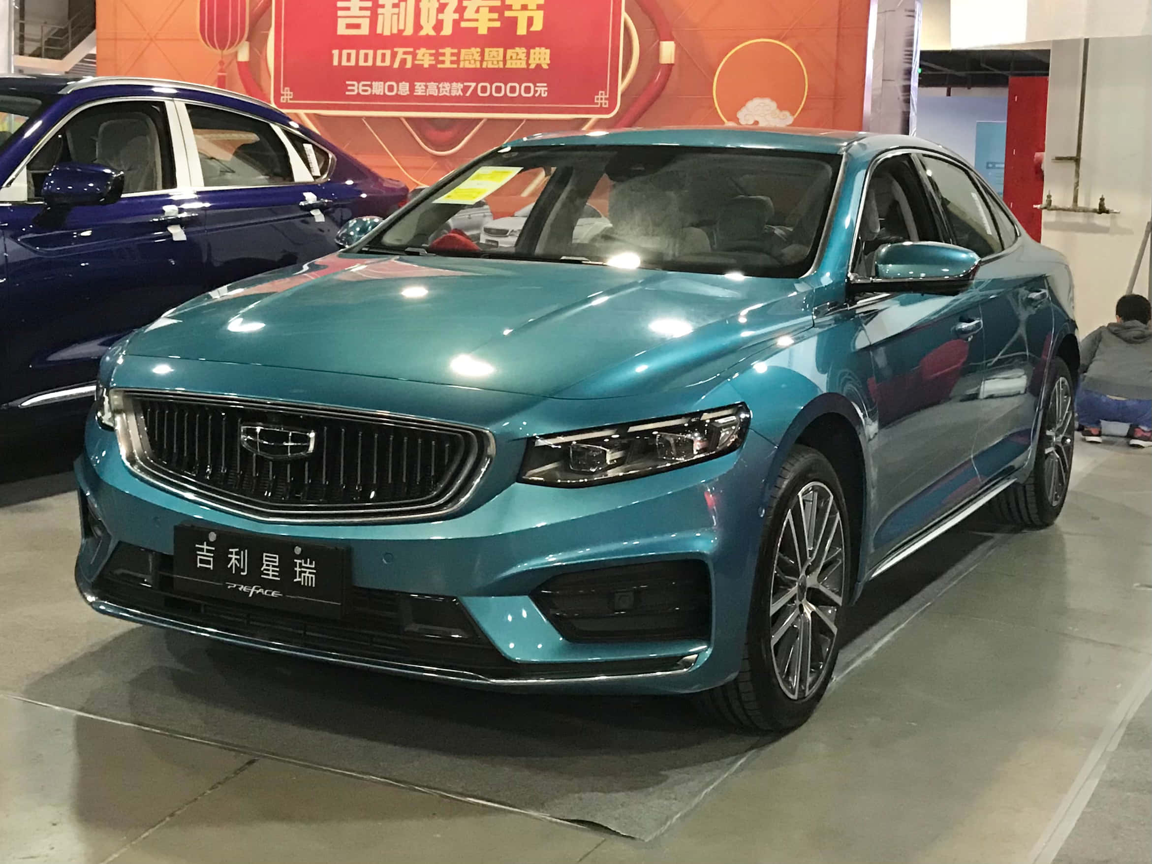 Geely Preface 桌布 壁紙 — 一輛閃亮的青綠色吉利Emgrand轎車在展廳燈光下閃耀，旁邊是一輛帶有宣傳橫幅的藍色汽車。