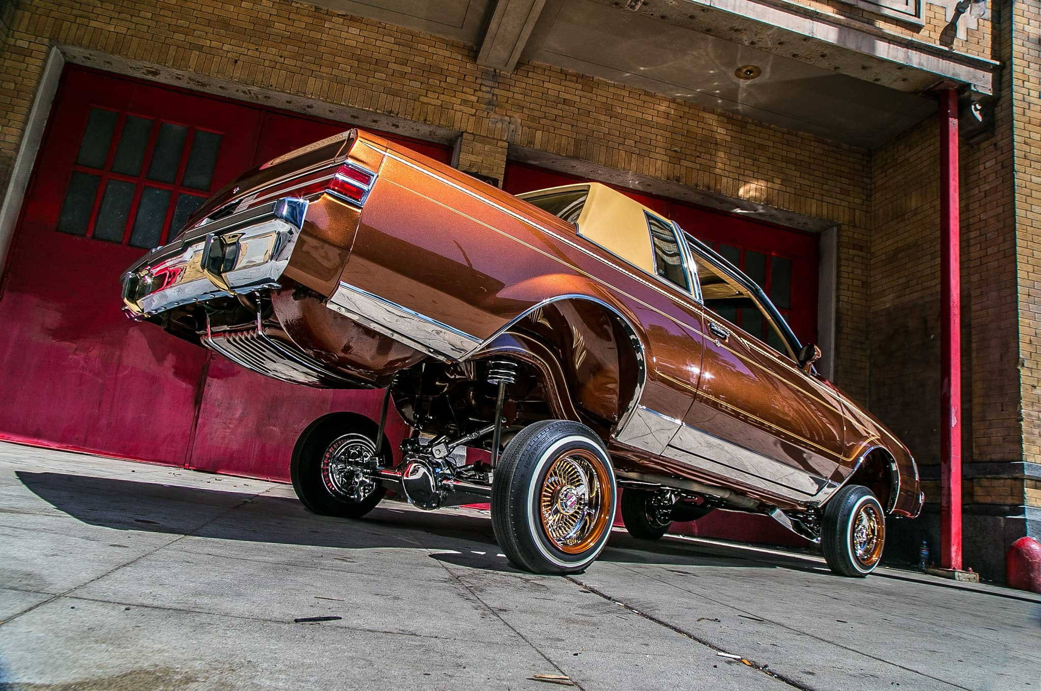 Lowrider 桌布 壁紙 — 一輛閃亮的青銅色定制低底盤汽車，帶有鍍鉻細節和金色邊緣輪圈，戲劇性地升起在混凝土車庫地板上。