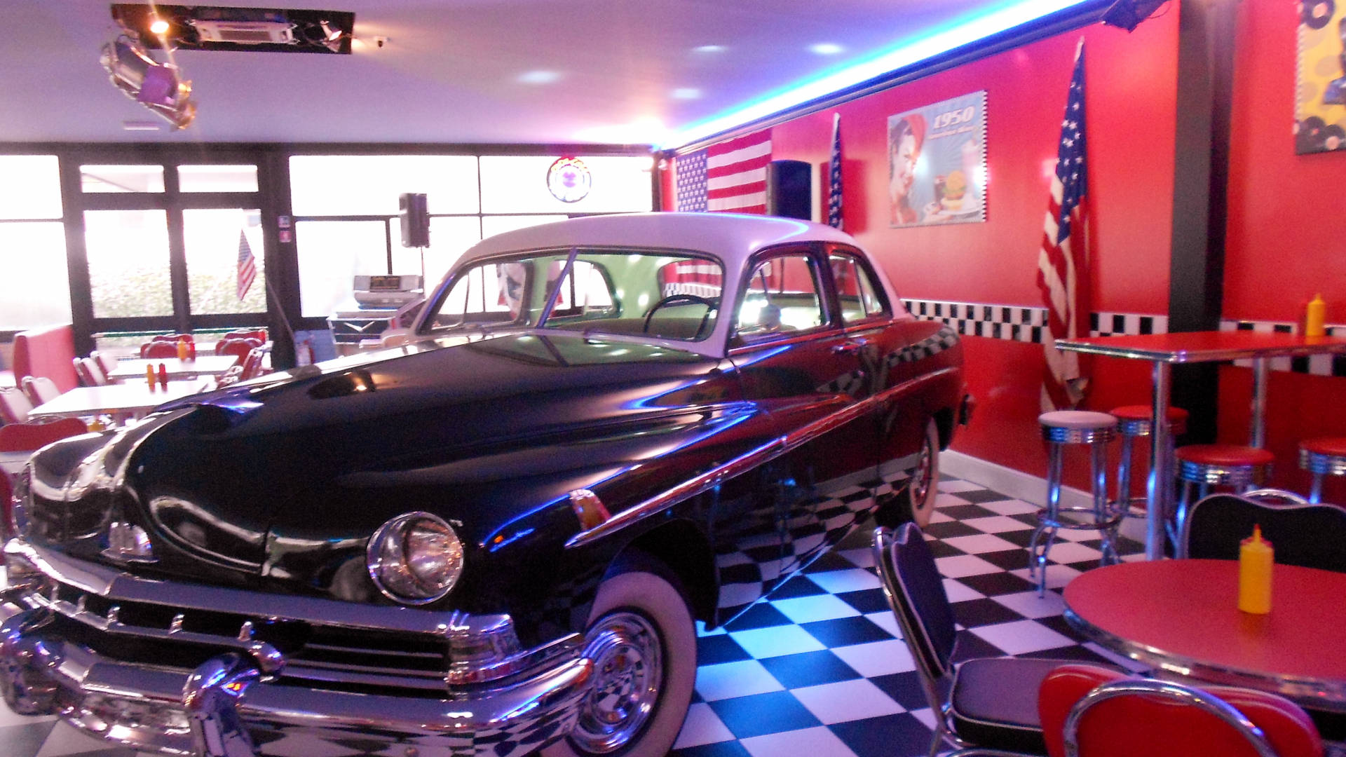 Diner 桌布 壁紙 — 一輛閃亮的黑色1950年代經典汽車在霓虹燈下，於一間設有紅色卡座和格子地板的復古餐廳中閃耀。