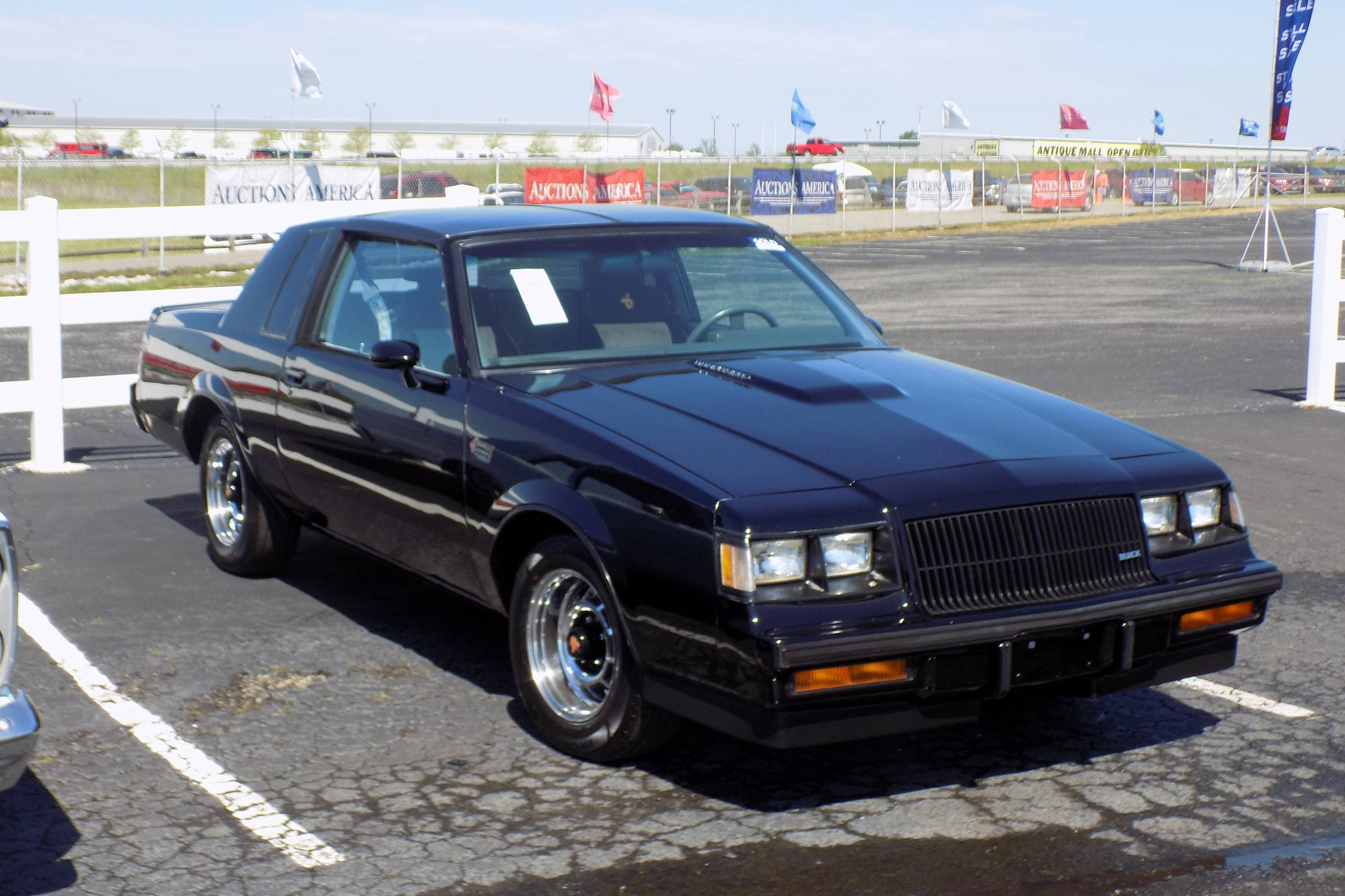 Buick 桌布 壁紙 — 一輛閃亮的黑色1980年代別克Grand National肌肉車停放在陽光明媚的戶外拍賣場，背景有白色圍欄和旗幟。