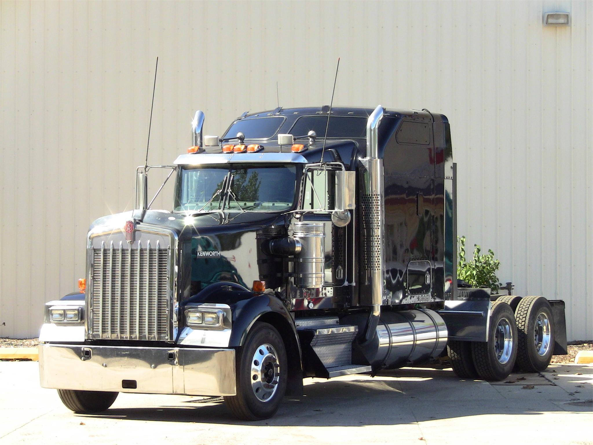 Kenworth W900 桌布 壁紙 — 一輛閃亮的黑色Kenworth半掛卡車，帶有鍍鉻裝飾，停在米色金屬建築前，陽光明媚。
