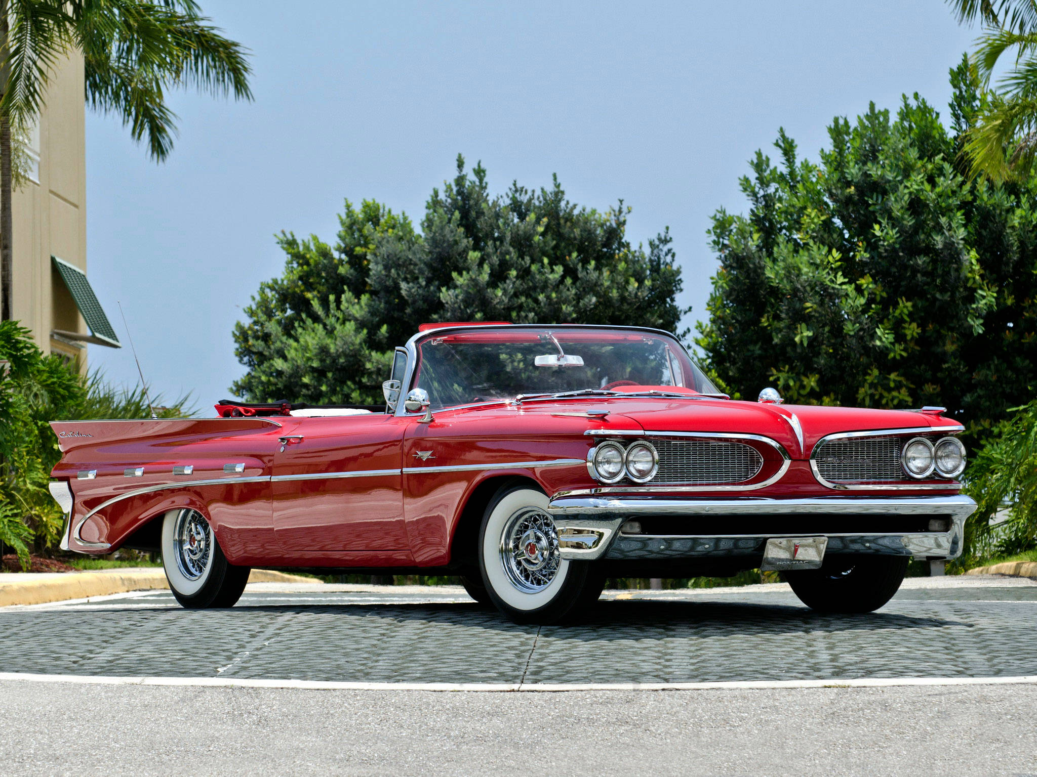 Pontiac 桌布 壁紙 — 一輛閃亮紅色1950年代敞篷車，帶有鍍鉻裝飾和白色側壁輪胎，停放在鋪砌的停車場上，背景是晴朗藍天、棕櫚樹和綠色灌木。