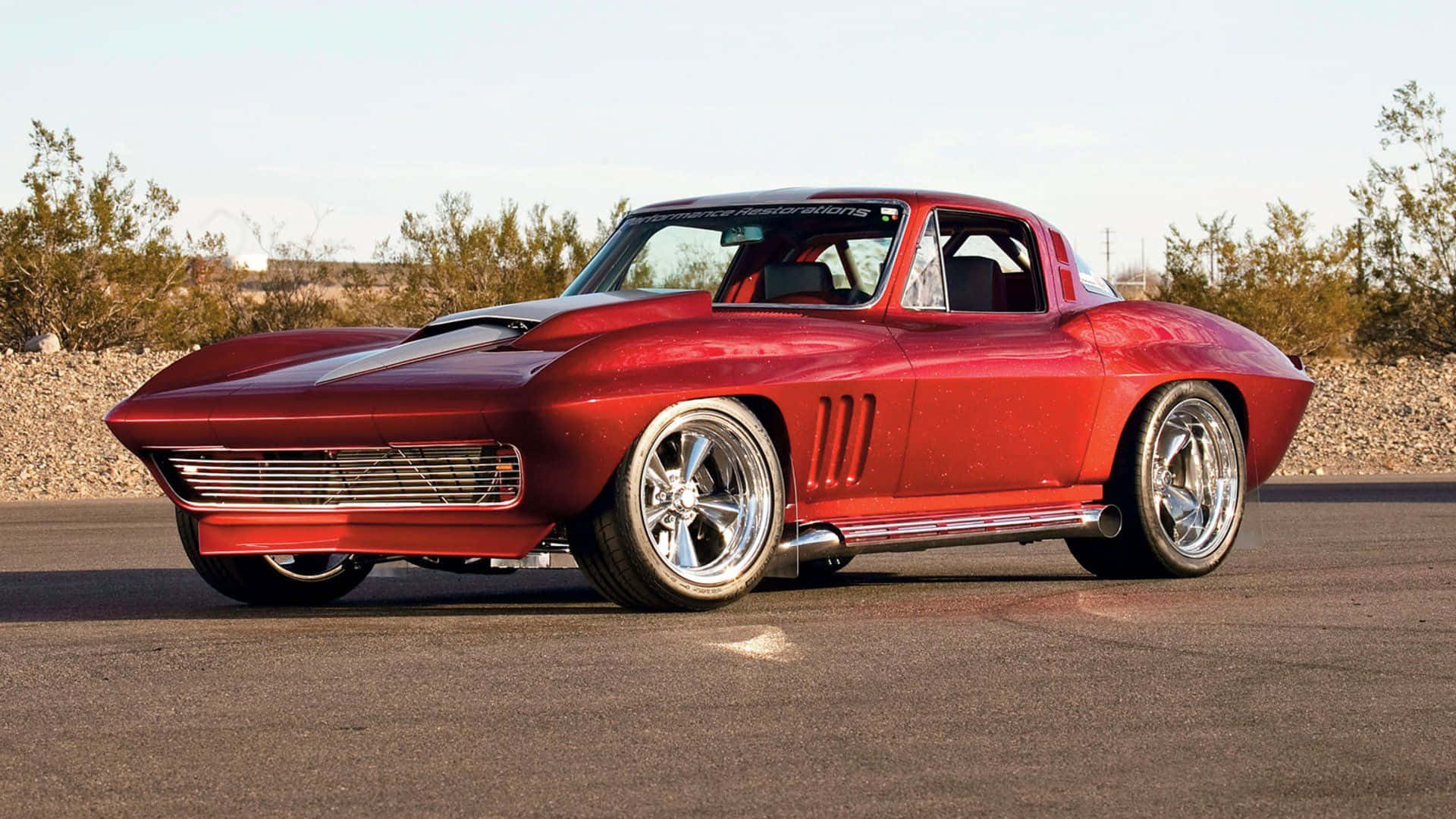 corvette 桌布 壁紙 — 一輛閃亮紅色經典肌肉車配備鉻合金輪圈，在明亮日光下停泊在瀝青路上。