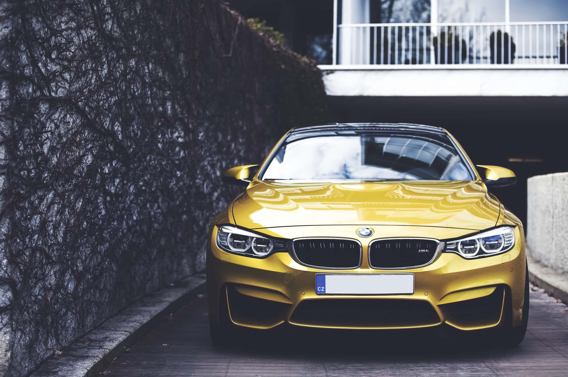 Bmw Car 壁紙 — 一輛閃亮金色BMW M4轎跑車停放在白色陽台下、深色長春藤牆旁的石徑上。