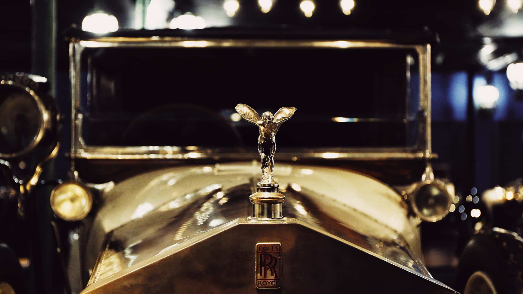 Luxury Brands 桌布 壁紙 — 一輛閃閃發光的復古勞斯萊斯，其標誌性的銀色歡慶女神引擎蓋飾物在溫暖的環境燈光下熠熠生輝。