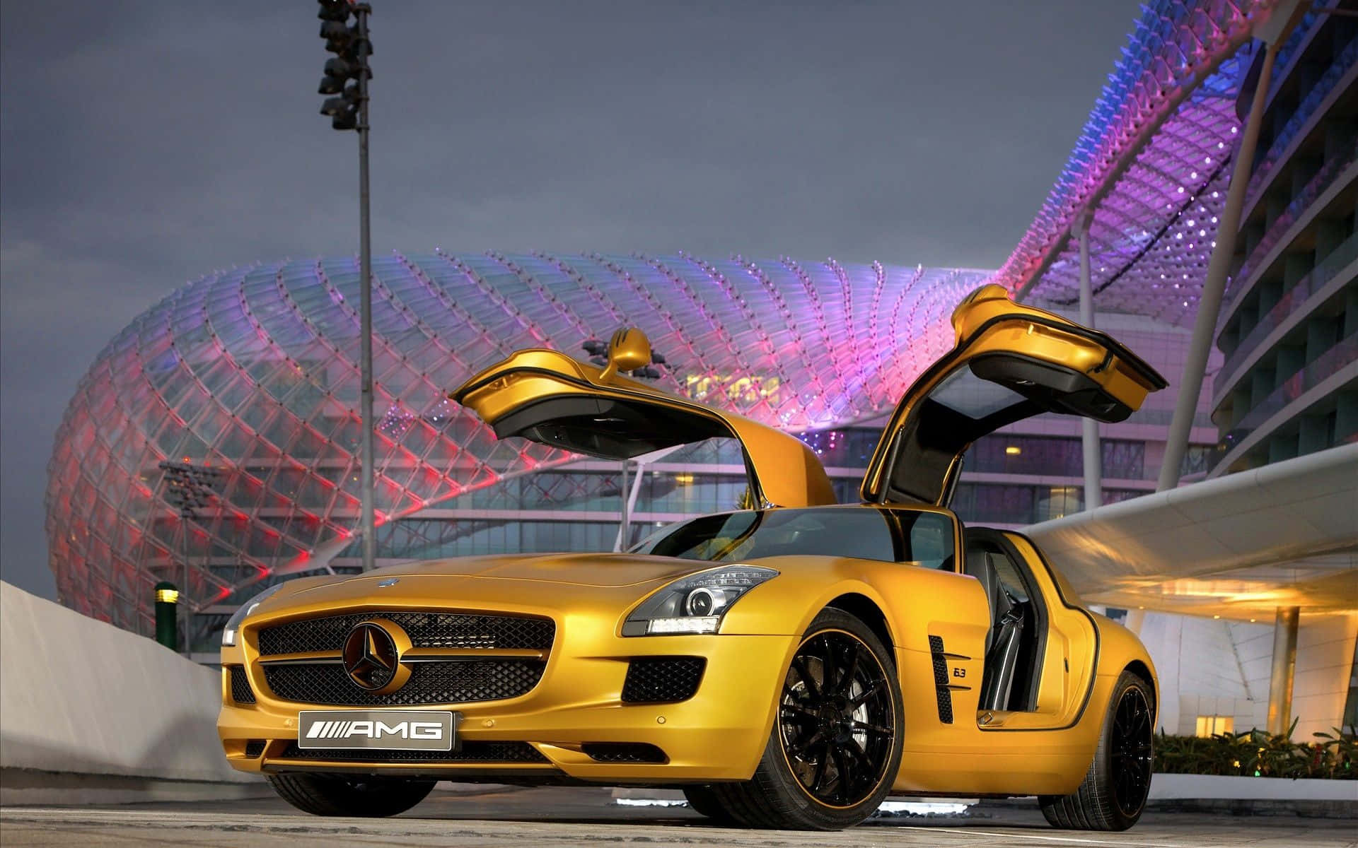 Gold Cars 桌布 壁紙 — 一輛閃閃發光的金色 Mercedes-AMG SLS 跑車，鷗翼車門打開，停放在黃昏時分一個未來感十足的發光圓頂結構前。
