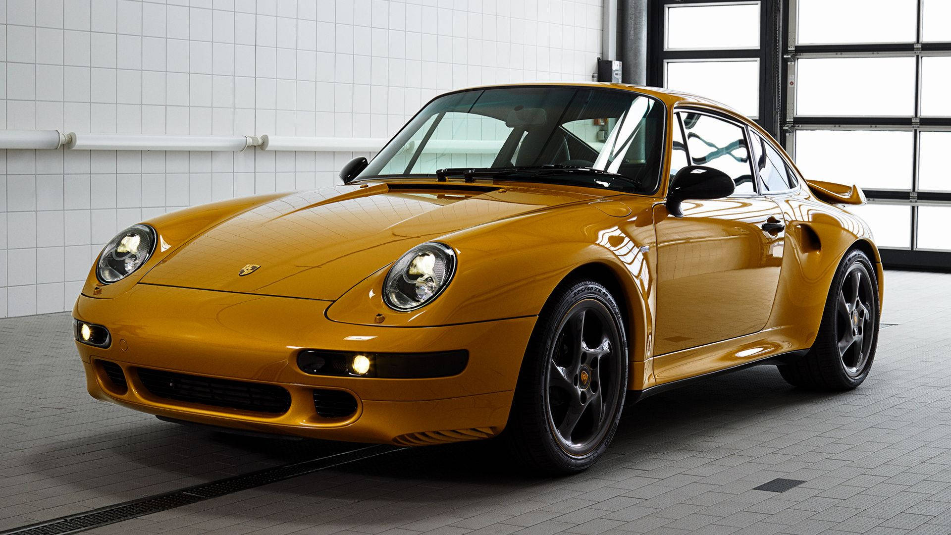 保時捷 911 桌布 壁紙 — 一輛閃閃發光的金色保時捷911 Turbo停在明亮的瓷磚車庫中，大窗戶將柔和的光線投射在其拋光的曲線上。