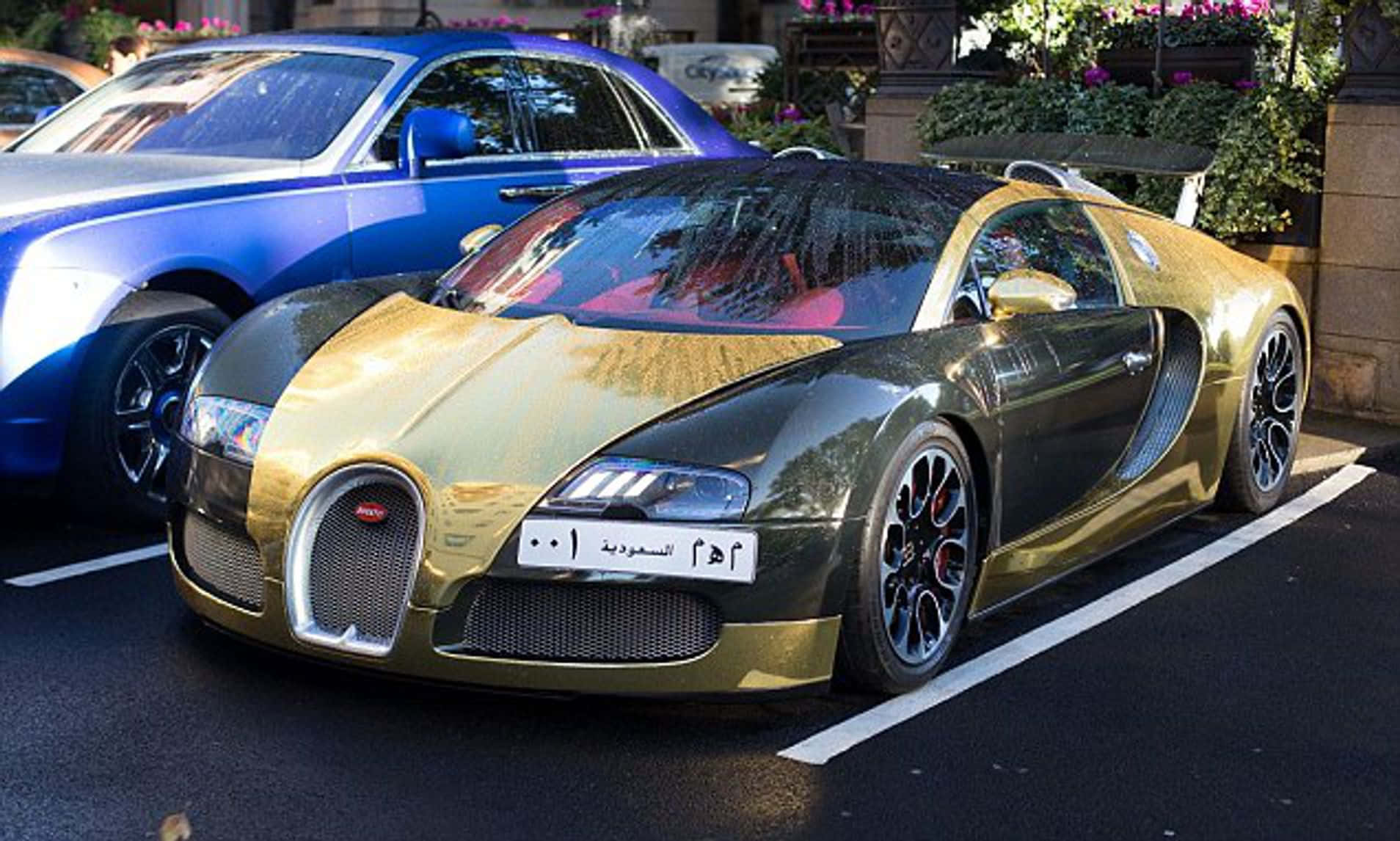 布加迪 桌布 壁紙 — 一輛閃閃發光的金色和黑色Bugatti Veyron停放在豪華城市停車場，旁邊有一輛藍色汽車，背景有綠化植物。