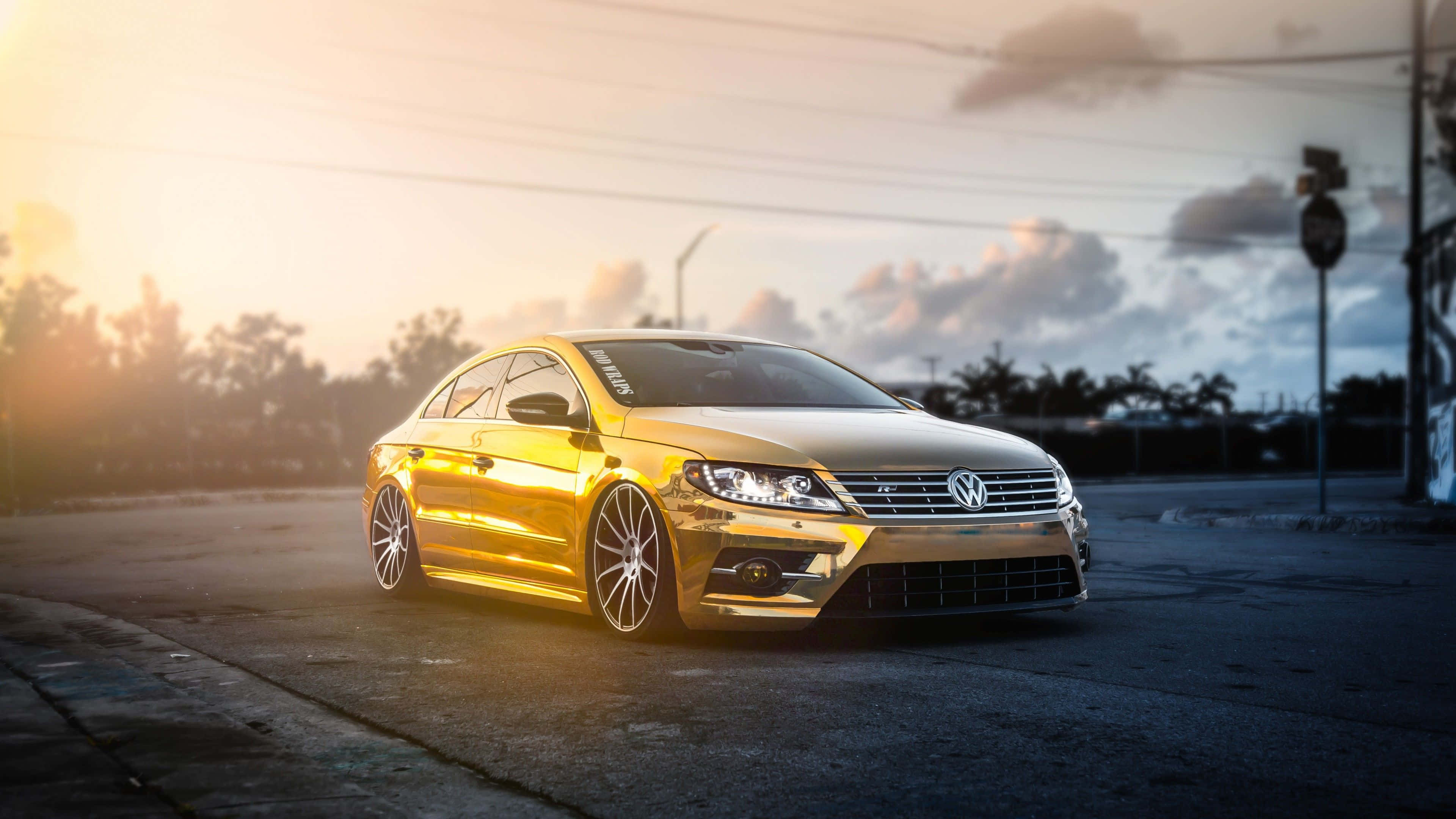 Gold Cars 桌布 壁紙 — 一輛閃閃發光的金色福斯房車在日落時分停在瀝青路上，帶有戲劇性的背光和陰天。