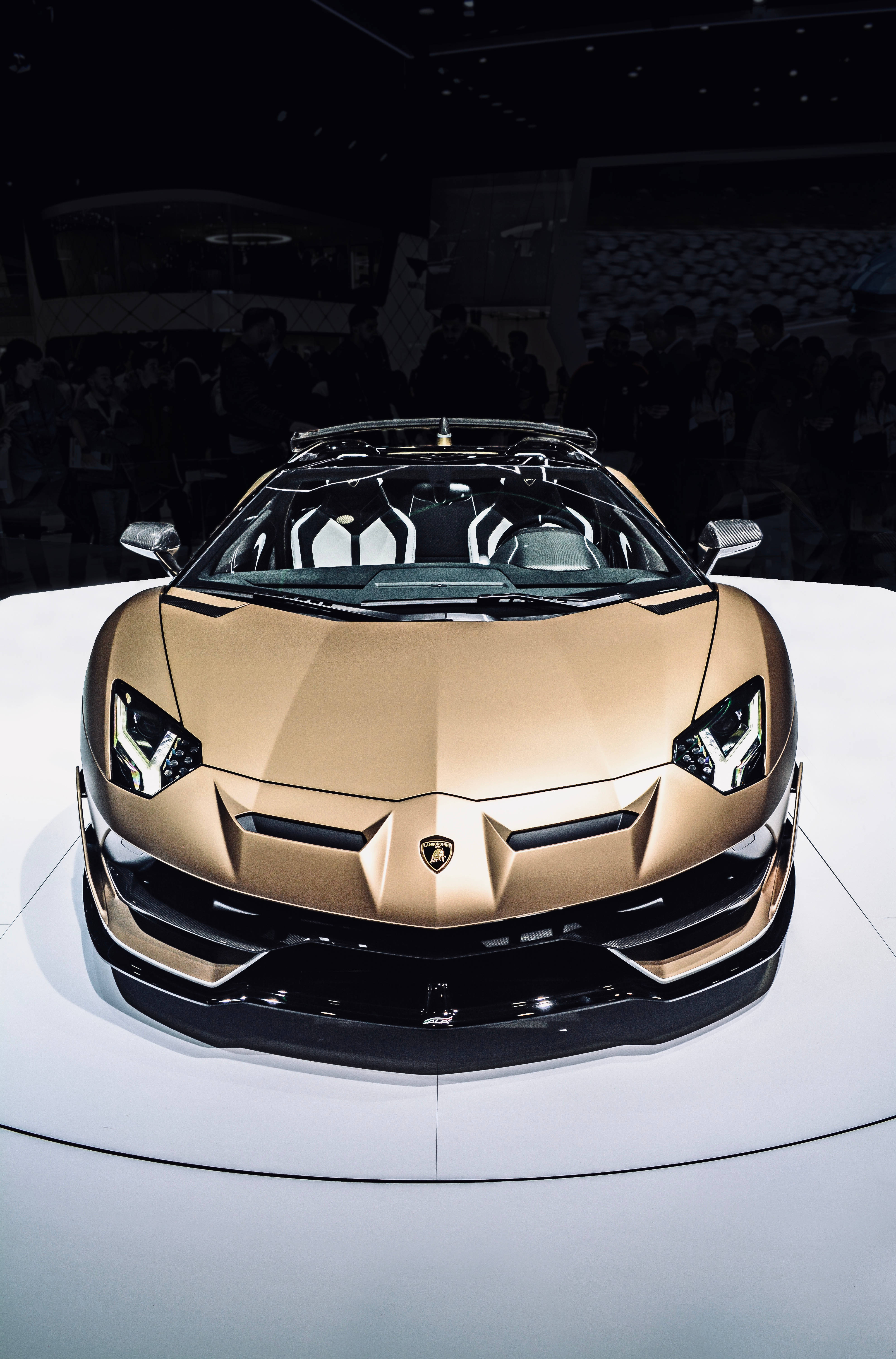 跑車 桌布 壁紙 — 一輛閃閃發光的金色藍寶堅尼 Aventador Roadster 在昏暗的車展中，白色平台上，背景有模糊的觀眾。