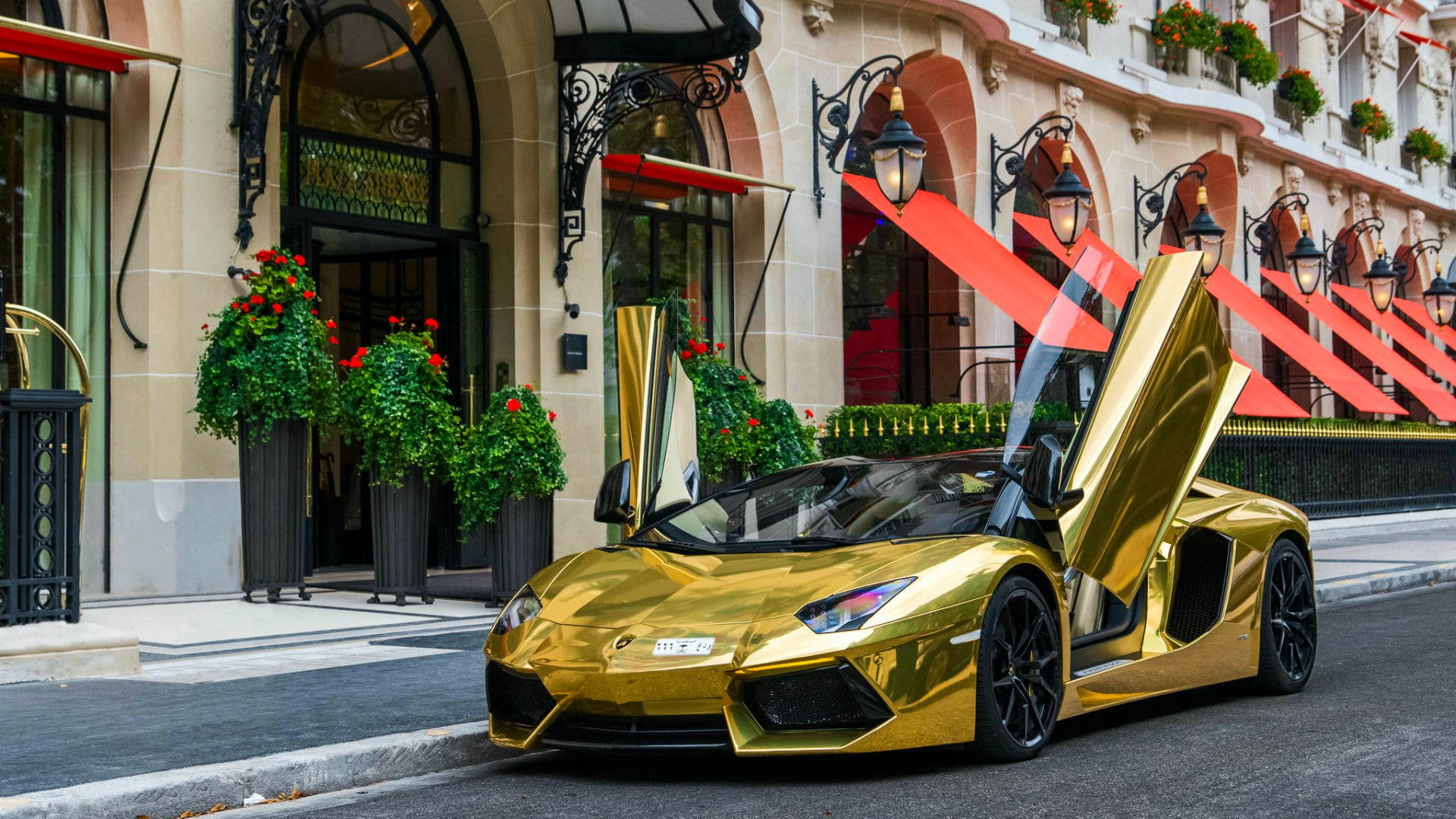 金色動態 桌布 壁紙 — 一輛閃閃發光的金色藍寶堅尼，剪刀門打開，停在時尚的巴黎街道上，街道兩旁有紅色遮陽篷和盆栽花卉。