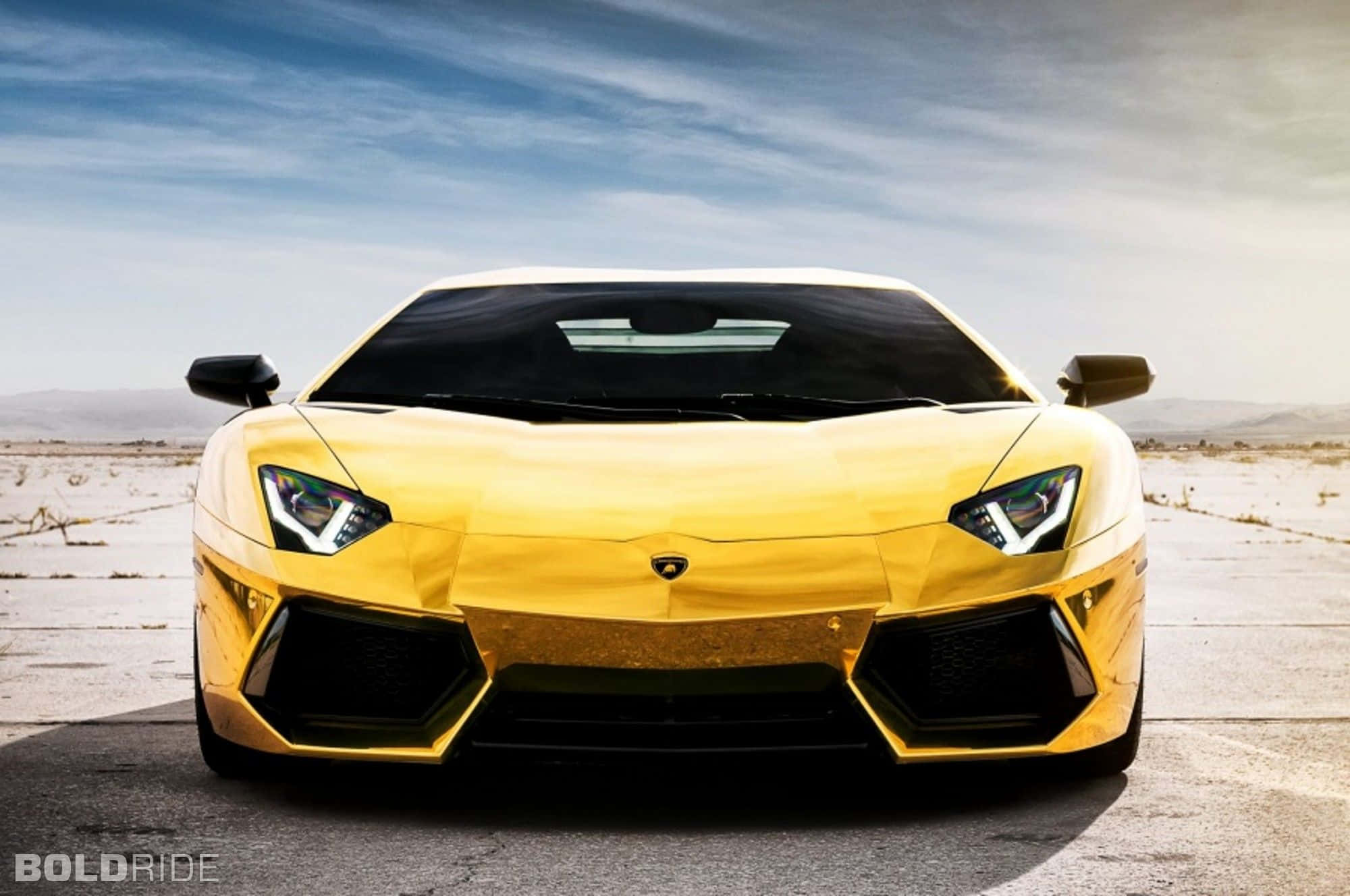 Gold Cars 桌布 壁紙 — 一輛閃閃發光的金色藍寶堅尼Aventador停泊在龜裂的沙漠道路上，廣闊的藍天中飄著縷縷輕雲。