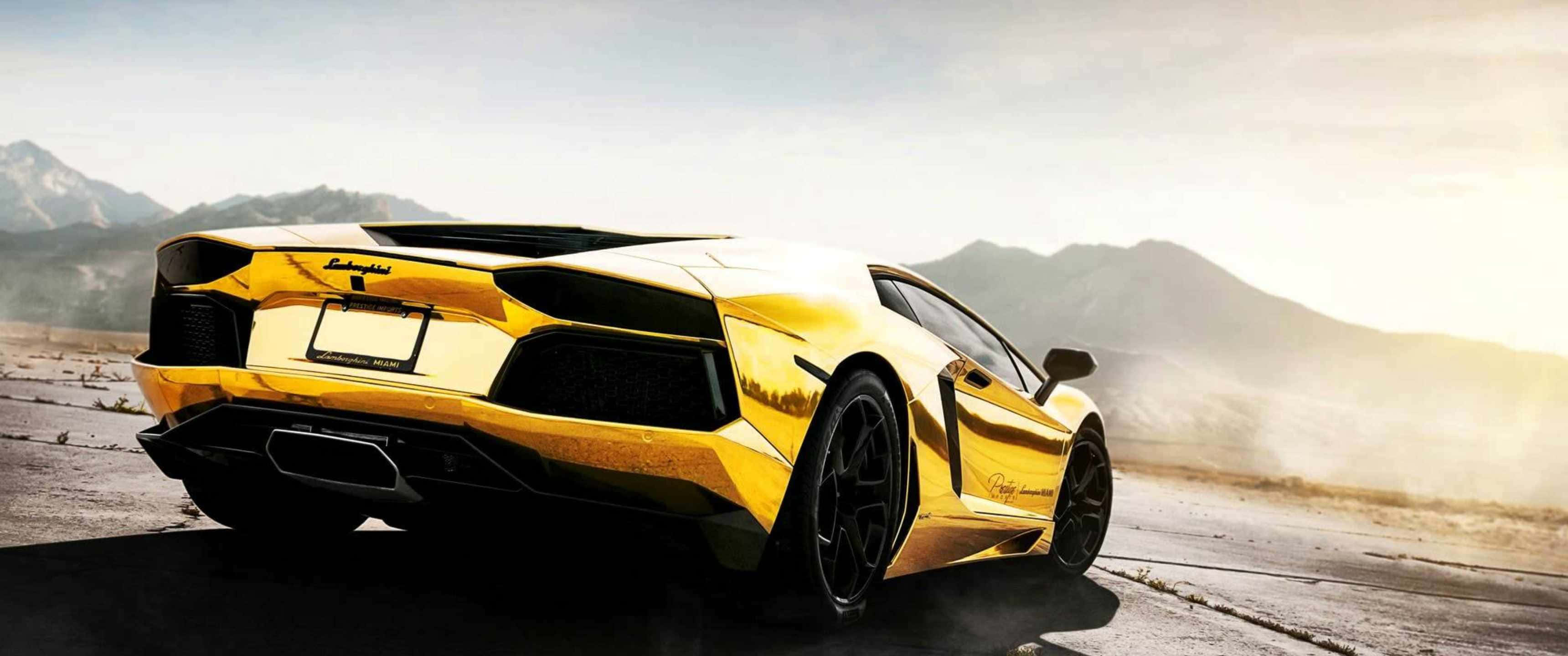 3440x1440p Lamborghini 桌布 壁紙 — 一輛閃閃發光的金色藍寶堅尼Aventador在沙漠公路上奔馳，背景是山脈，天空朦朧。