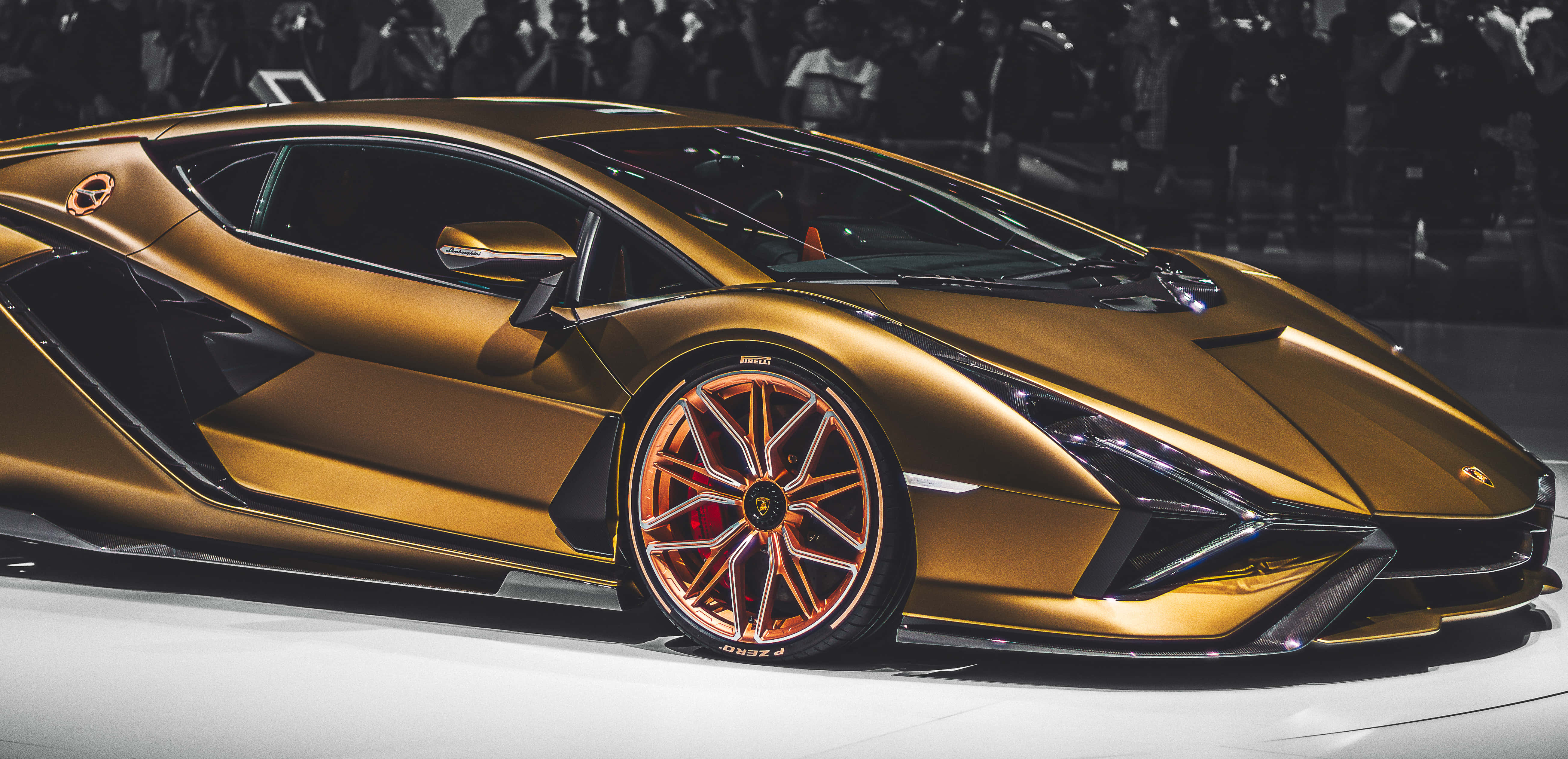Gold Cars 桌布 壁紙 — 一輛閃閃發光的金色藍寶堅尼跑車搭配橙色輪胎，在白色平台上於明亮燈光下展示，背景是模糊的人群。