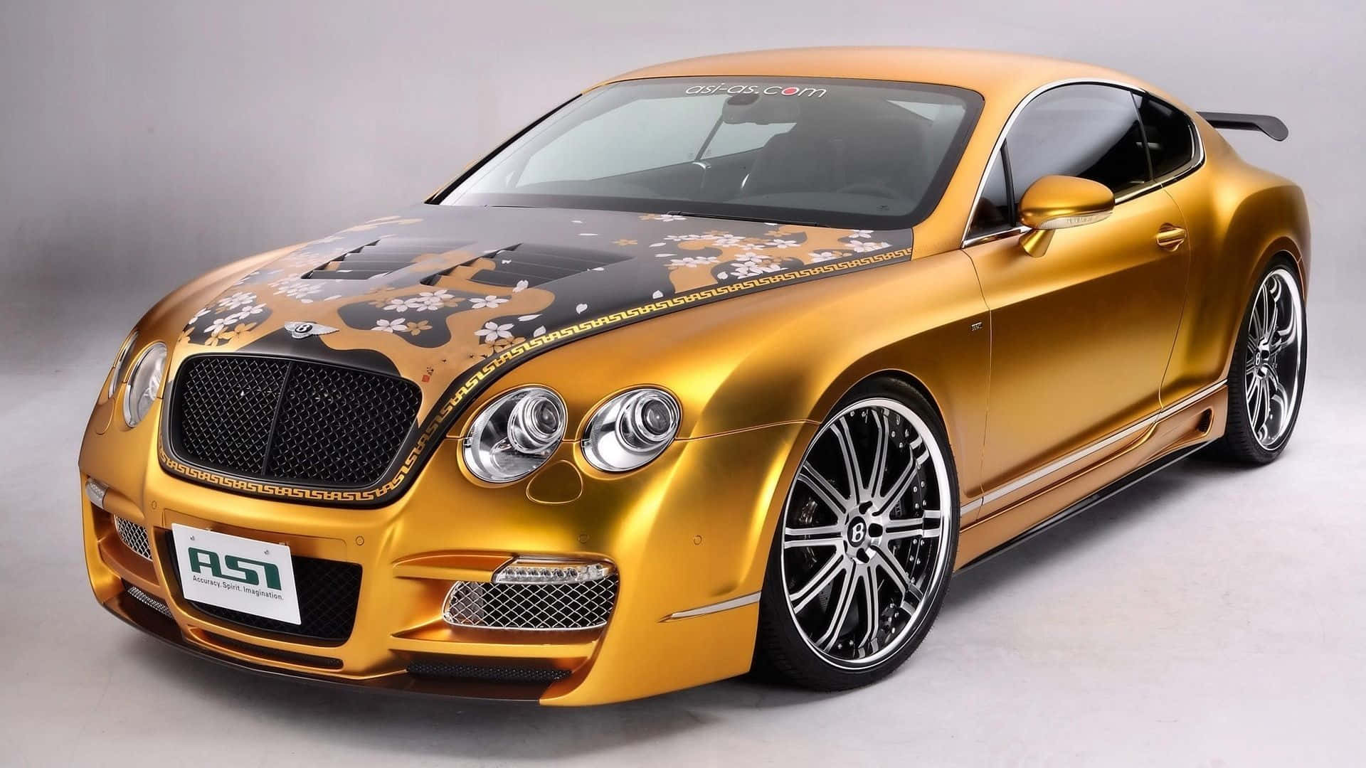 Luxury 桌布 壁紙 — 一輛閃閃發光的金色賓利 Continental GT，帶有黑色花卉引擎蓋藝術和定制輪圈，在攝影棚中擺拍。