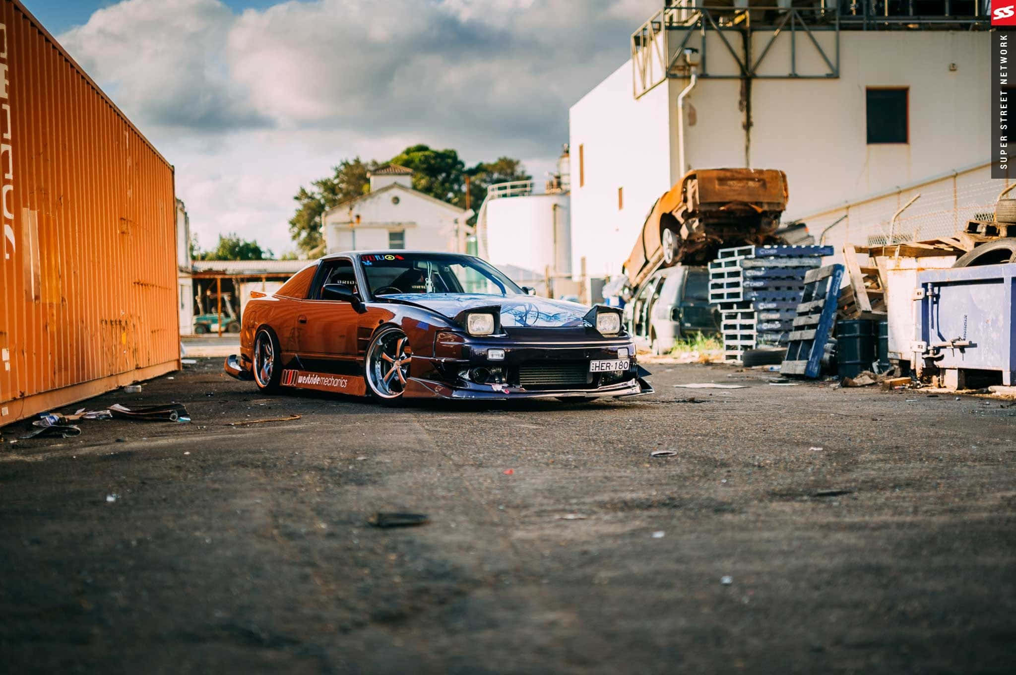 Nissan 180sx 桌布 壁紙 — 一輛降低的橙色和藍色跑車停放在粗糙的工業場地，旁邊是貨櫃，天空多雲。
