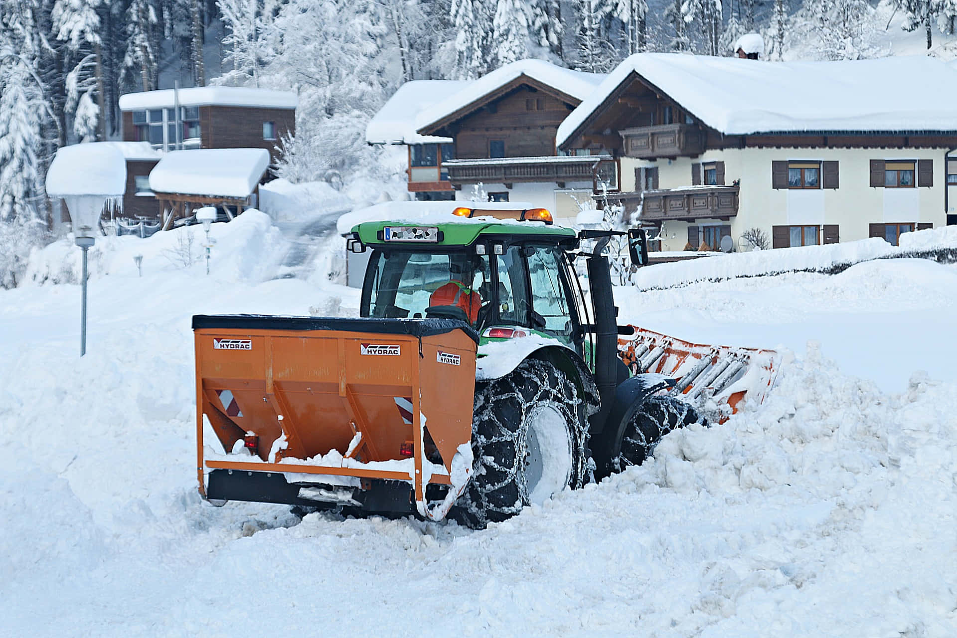 Snowplow 桌布 壁紙 — 一輛除雪拖拉機正在清理一條寧靜的村莊街道上厚厚的積雪，街道兩旁是白雪覆蓋的小木屋和松樹。