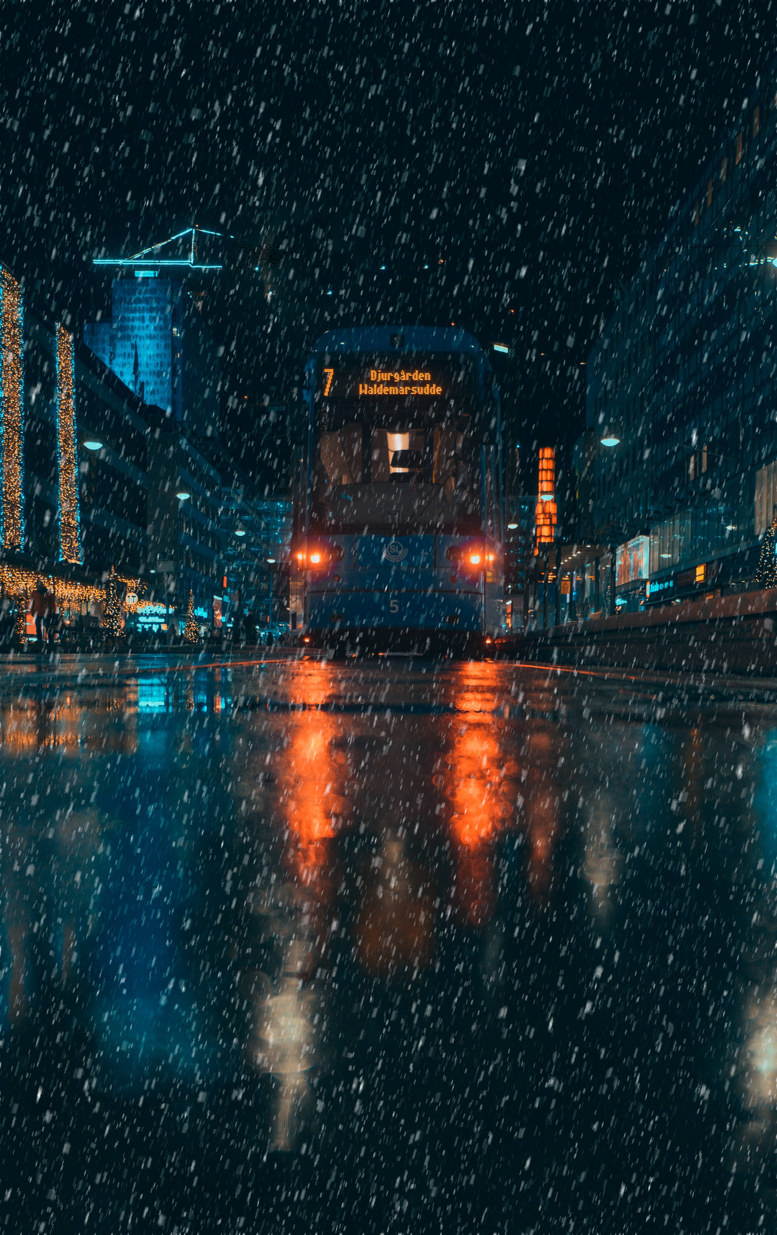 下雨 桌布 壁紙 — 一輛電車在雪夜的城市中滑行，其紅燈在落雪下映照在濕漉漉的路面上。