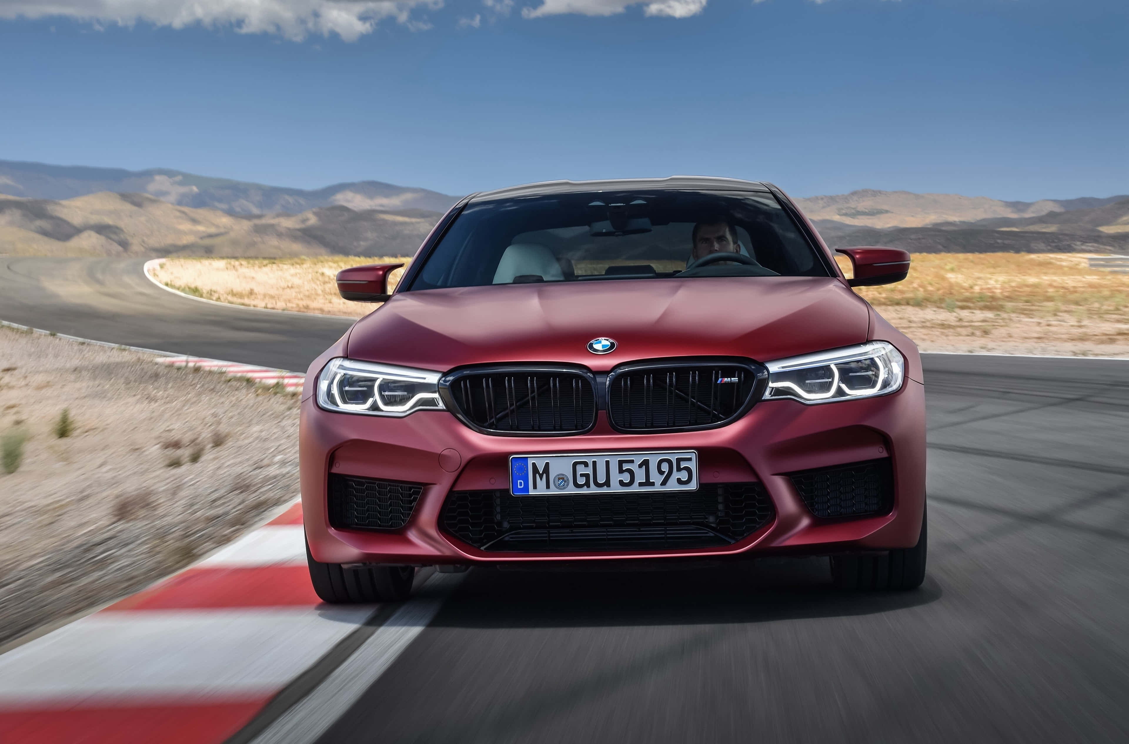 桌布 壁紙 — 一輛霧面紅色BMW M5在晴朗的藍天下，沿著沙漠賽道疾馳，呈現模糊的動感。