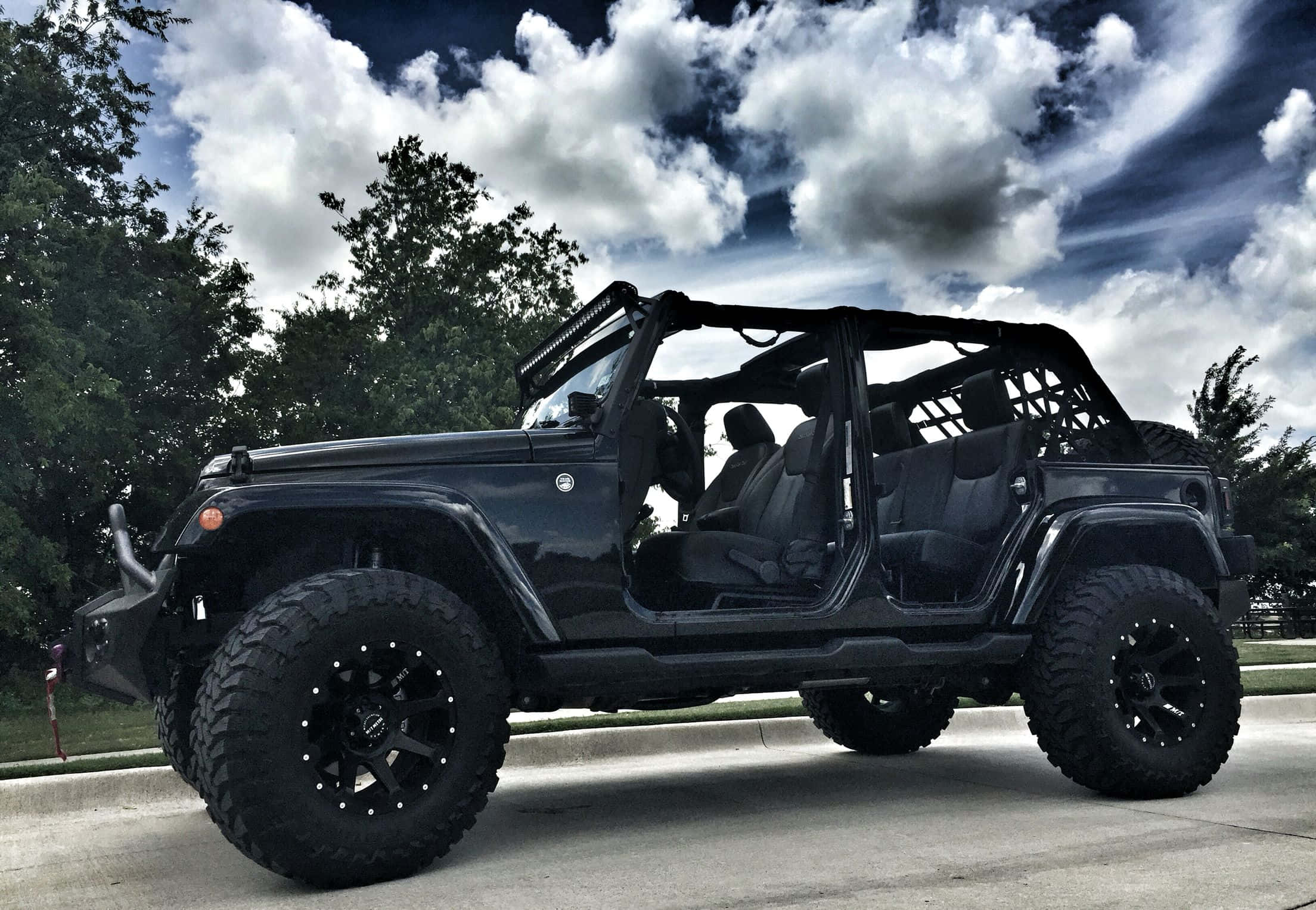 黑色吉普車 桌布 壁紙 — 一輛霧面黑色Jeep Wrangler，配備超大輪胎和打開的車門，停在戲劇性多雲天空下的混凝土車道上。