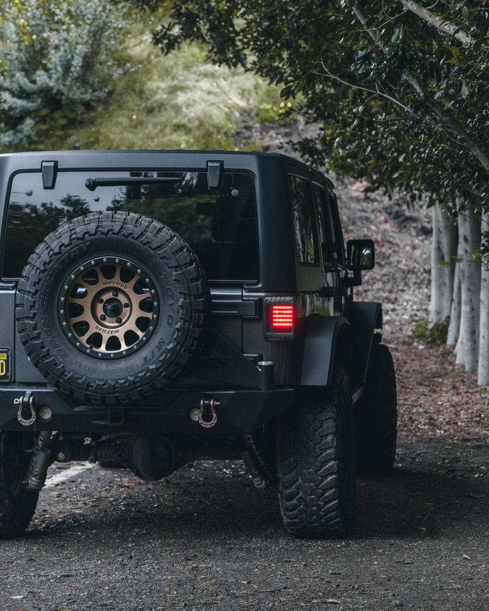 黑色吉普車 桌布 壁紙 — 一輛霧面黑色Jeep Wrangler，後方裝有備用輪胎，行駛在樹木成蔭的碎石路上。