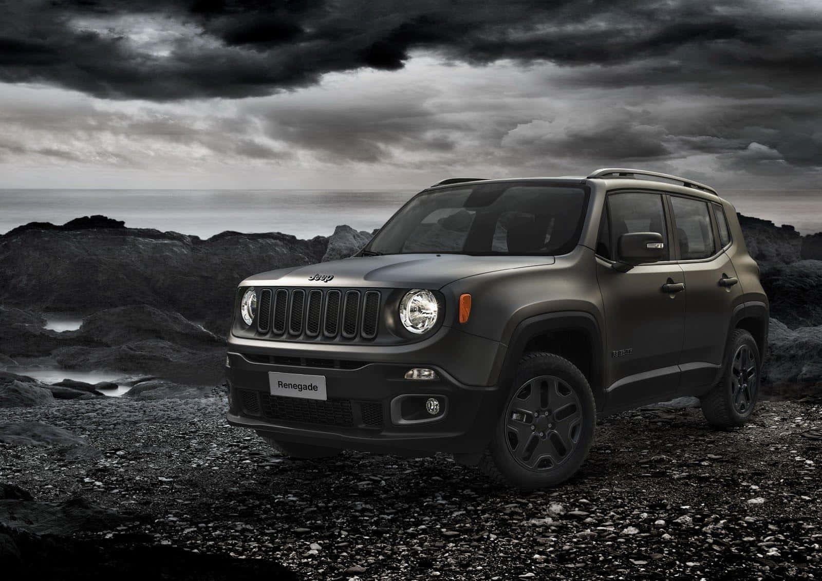 Jeep Renegade 桌布 壁紙 — 一輛霧面黑色的Jeep Renegade堅固地矗立在岩石海岸上，頭上是暴風雨前的灰色天空，海浪拍打著岸邊。