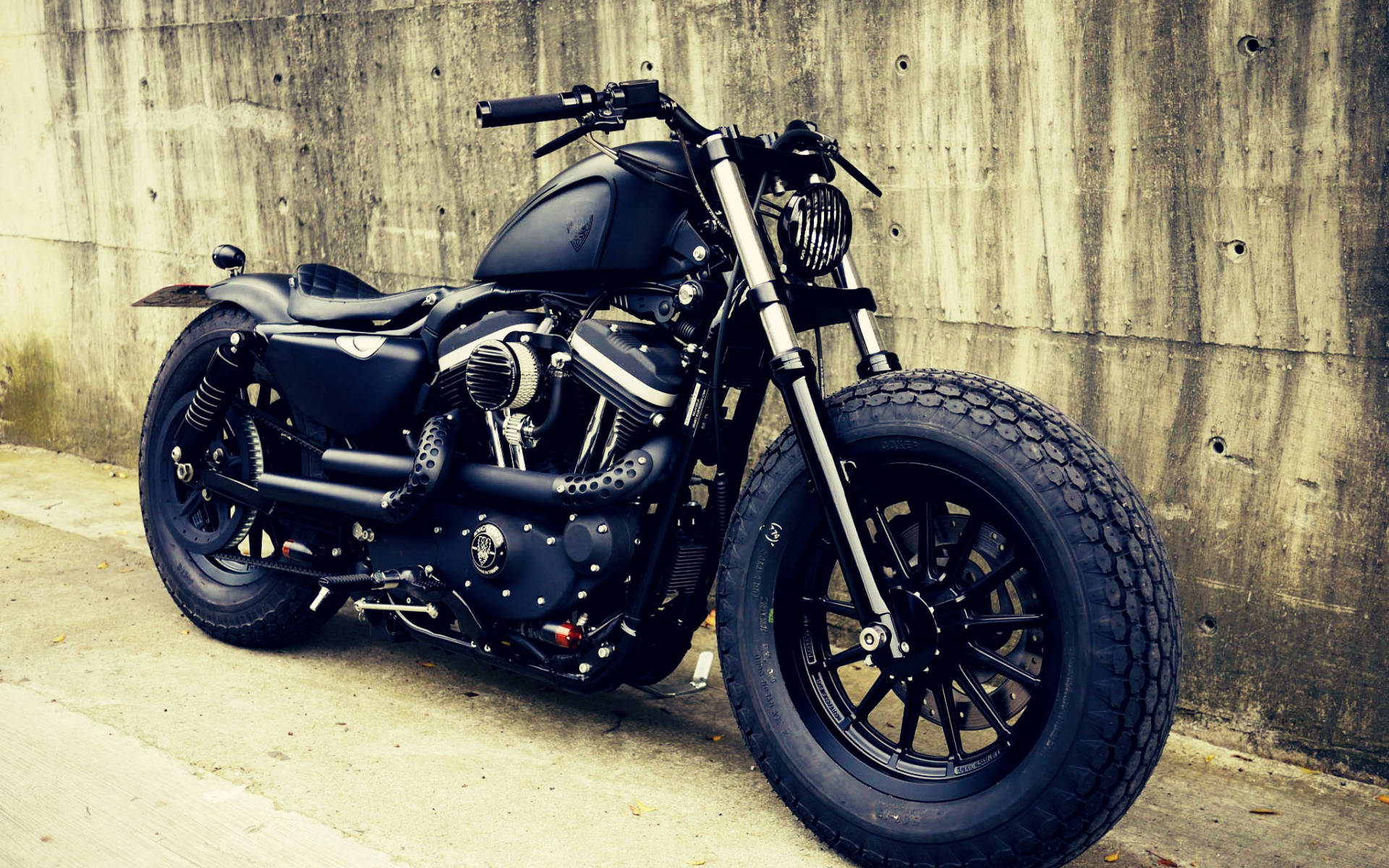 Bobber 桌布 壁紙 — 一輛霧面黑色訂製chopper機車，配備寬輪胎，靠在人行道上風化的混凝土牆邊。