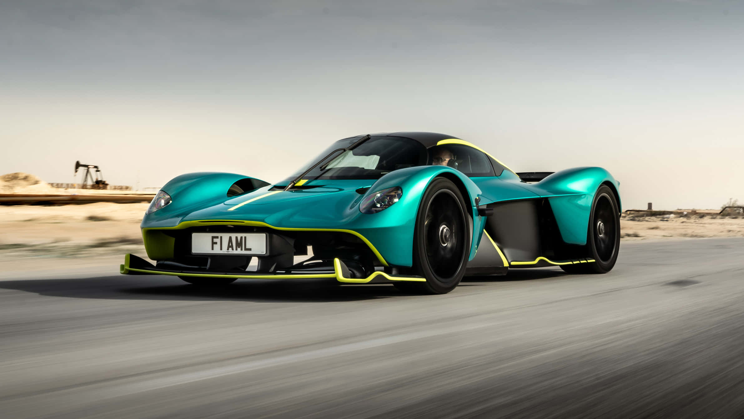 奧斯頓馬丁 桌布 壁紙 — 一輛青綠色和黃色的Aston Martin Valkyrie hypercar在沙漠公路上飛馳，背景有石油鑽井平台。