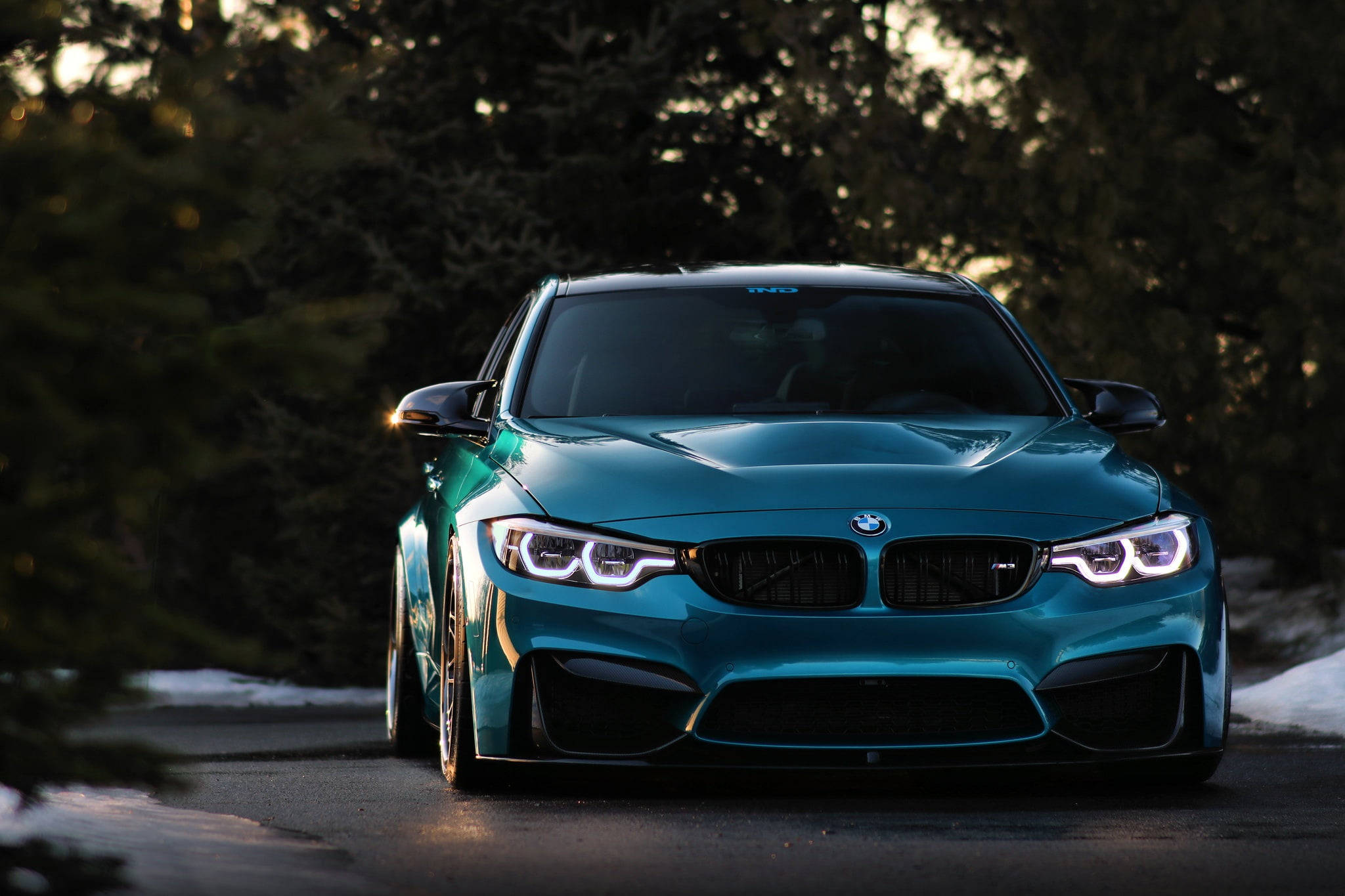 BMW 動圖 桌布 壁紙 — 一輛青綠色的BMW M4跑車在白雪覆蓋的森林道路上，在金黃色的夕陽光下閃閃發光。