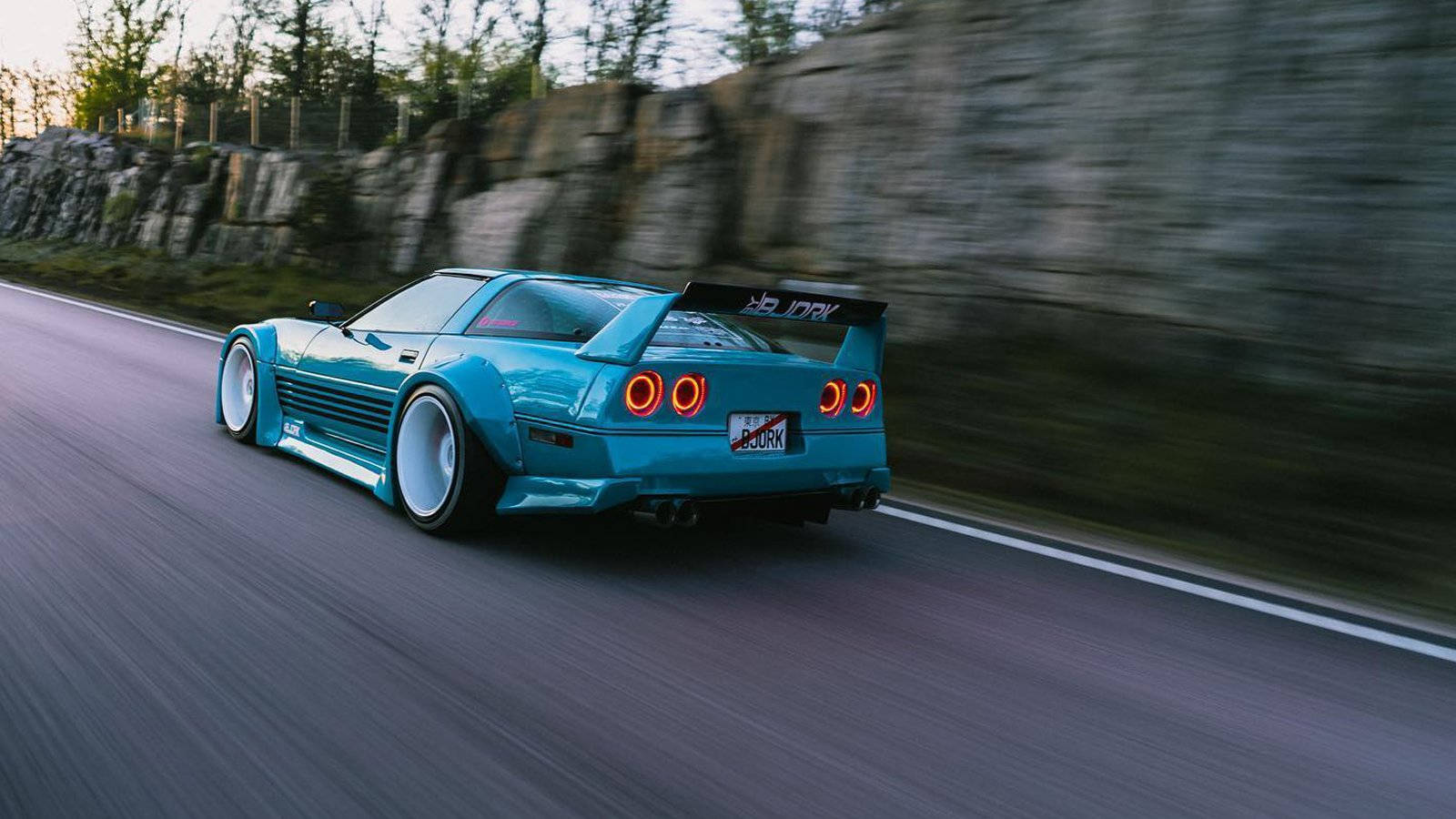 corvette 桌布 壁紙 — 一輛青綠色的改裝跑車在蜿蜒的道路上疾馳，背景是岩石峭壁，帶有動態模糊效果。