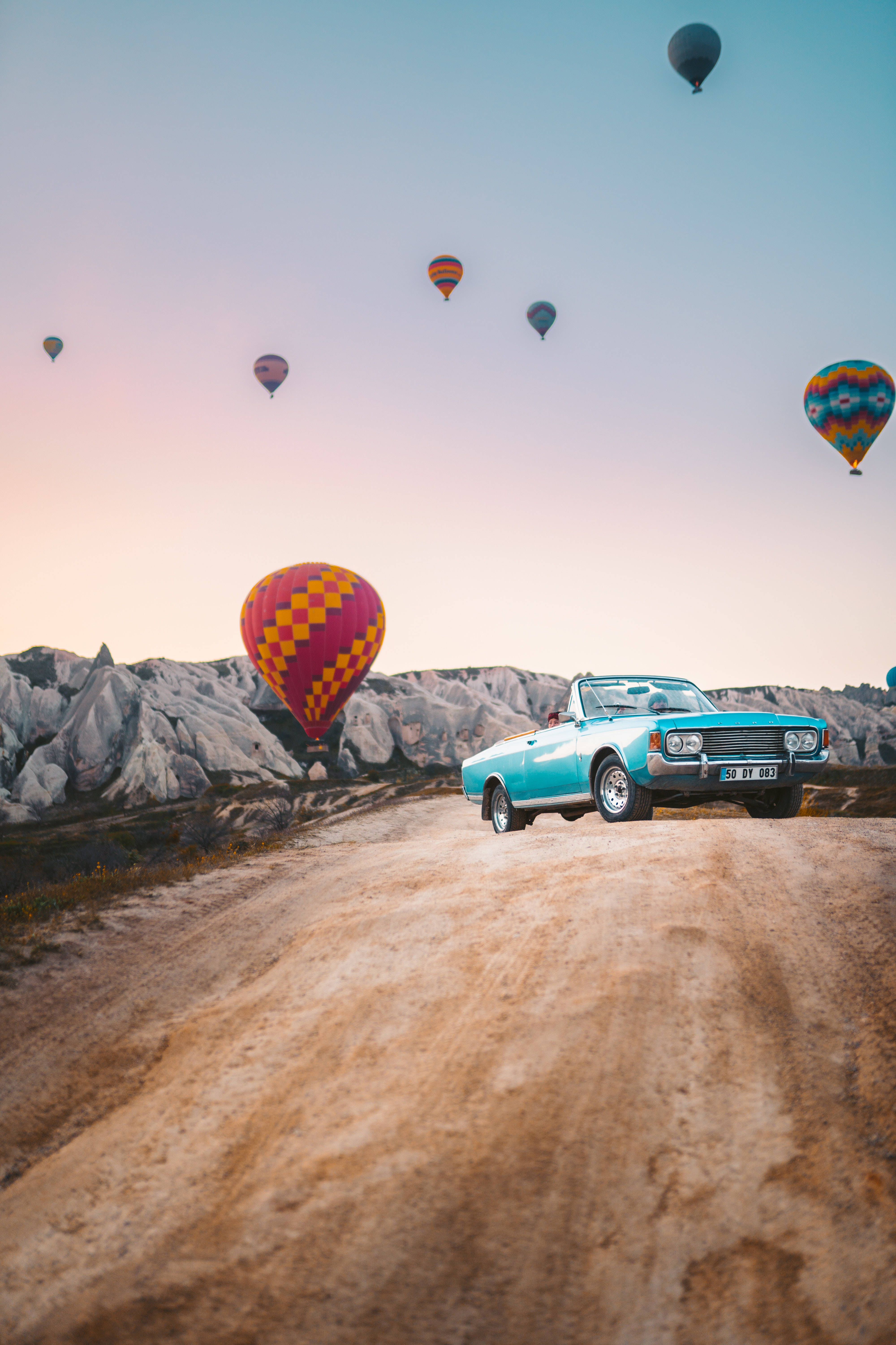 Cappadocia 桌布 壁紙 — 一輛青綠色的敞篷車在日落時分，行駛在一條塵土飛揚的道路上，下方是超現實的岩石地貌，上方飄浮著色彩繽紛的熱氣球。