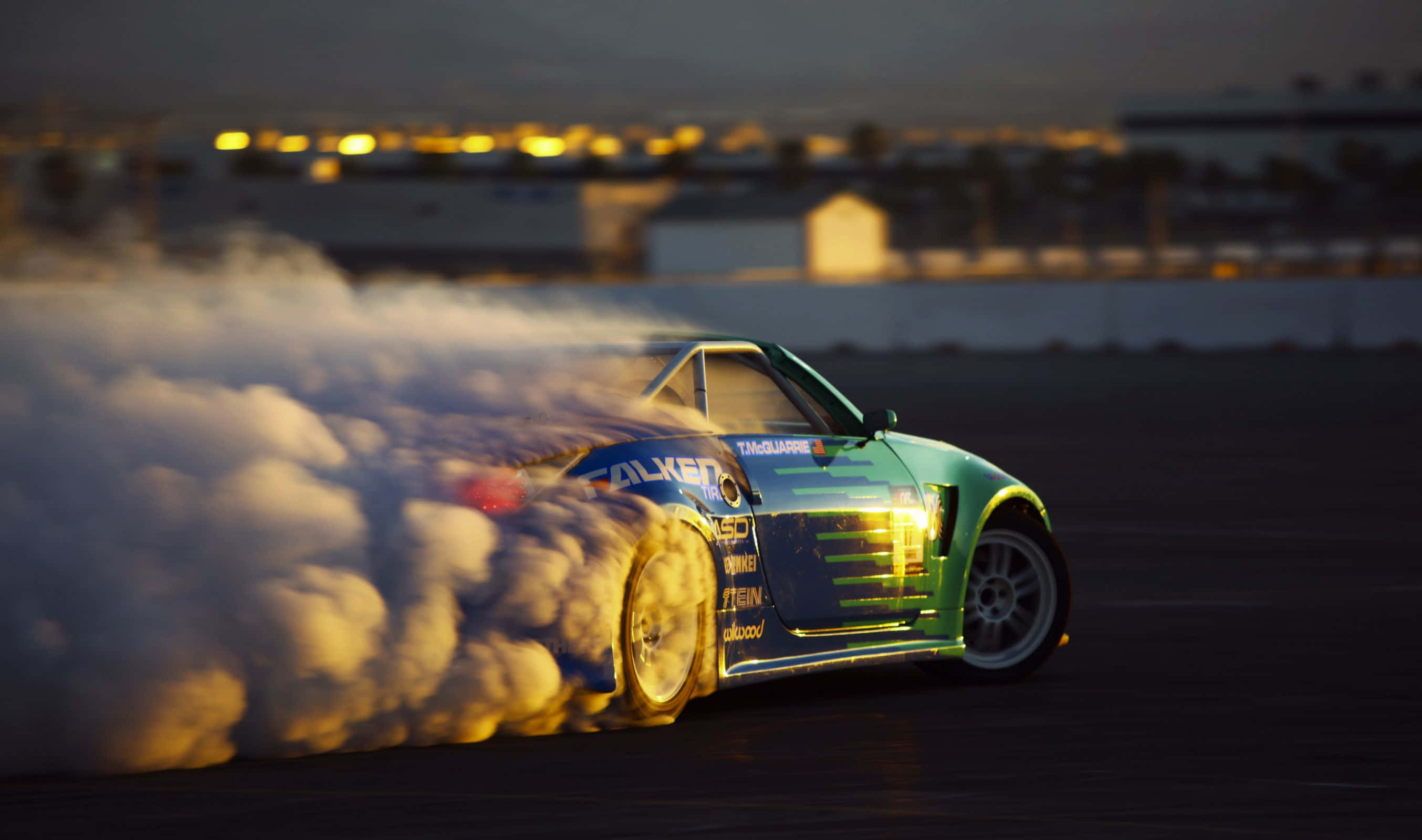 Racing 桌布 壁紙 — 一輛青綠色的漂移車在黃昏時分在黑暗的賽道上滑行，揚起濃煙，遠處的城市燈光閃爍。