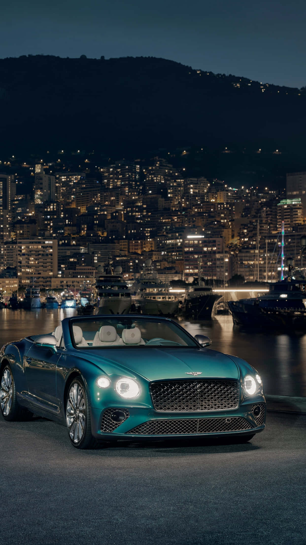 Bentley Continental Gt 桌布 壁紙 — 一輛青綠色賓利敞篷車在夜燈下閃閃發光，停泊在港口旁，背後的山丘上閃爍著城市景觀。