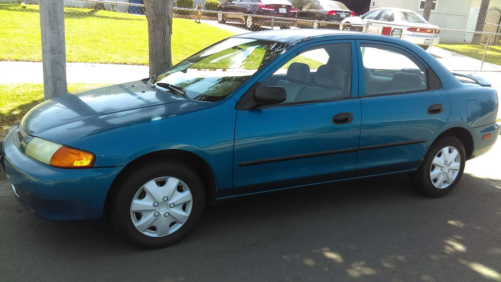 Mazda Protege 桌布 壁紙 — 一輛青綠色轎車停放在陽光明媚的街道上，背景有樹木和其他車輛。