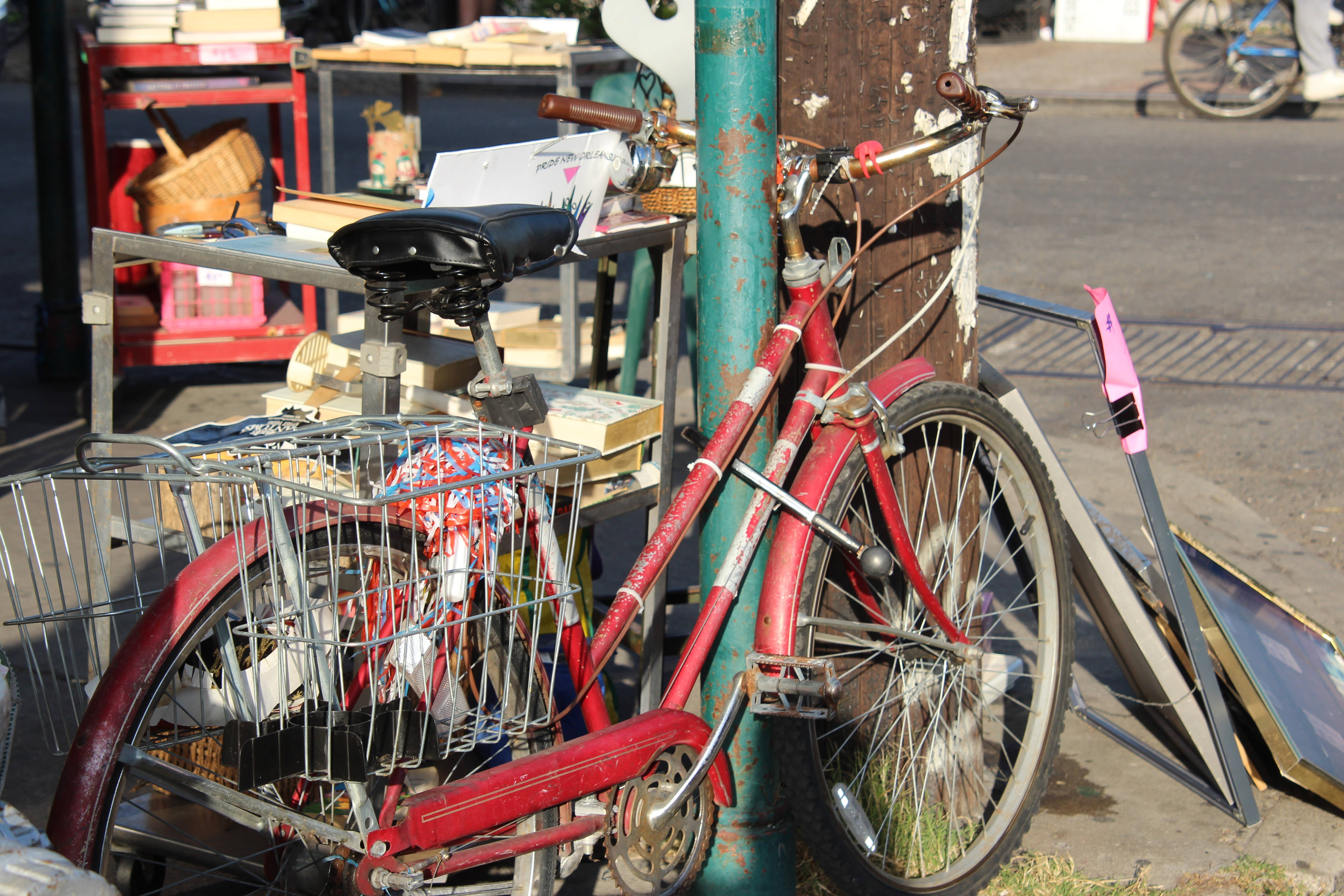 Bicycle 桌布 壁紙 — 一輛風化的紅色腳踏車靠在綠色柱子上，旁邊是雜亂的戶外攤位，在明亮的日光下。