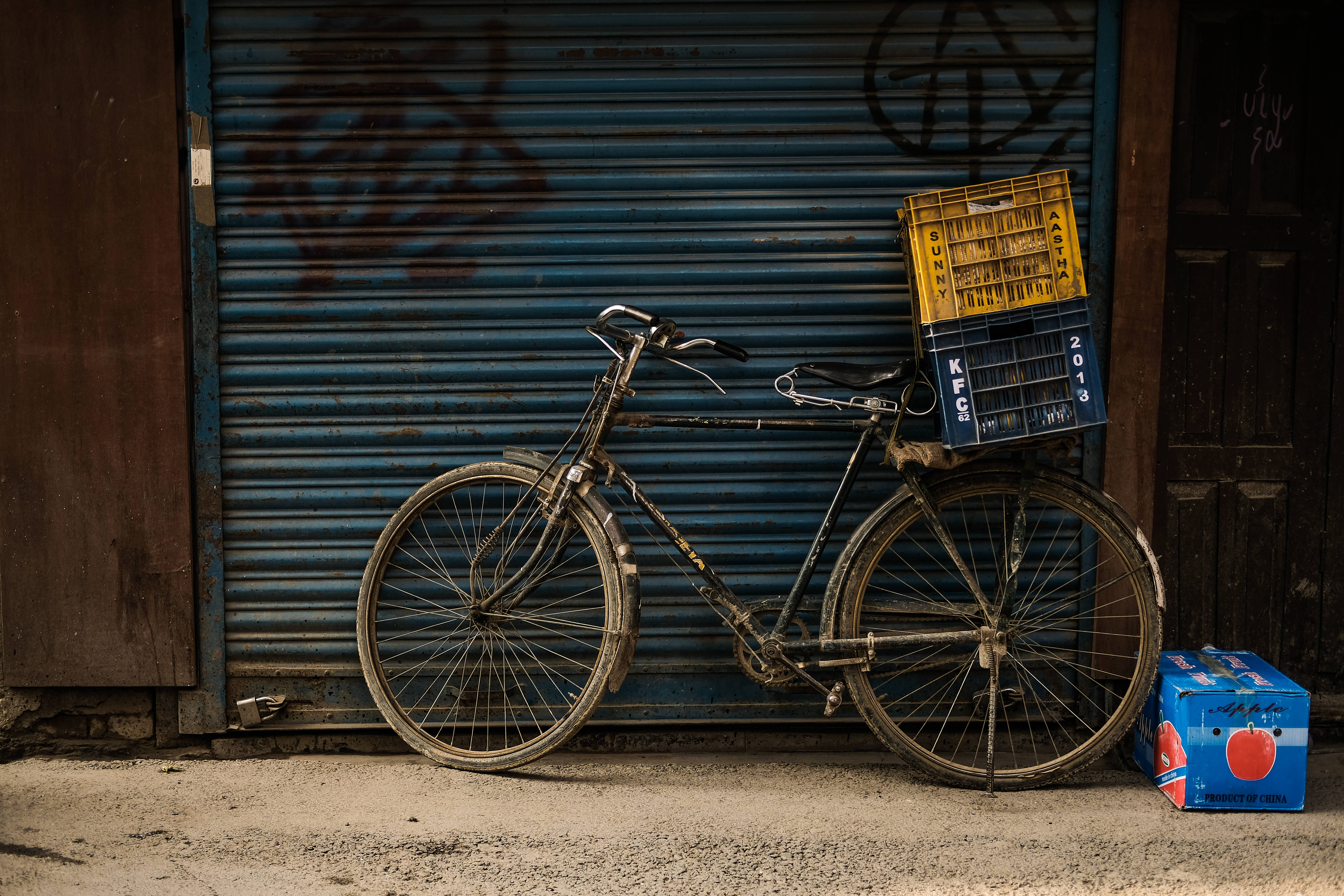 Nepal 桌布 壁紙 — 一輛飽經風霜的自行車，後座載著一個黃藍色的板條箱，靠在一扇佈滿塗鴉的藍色金屬百葉窗上。