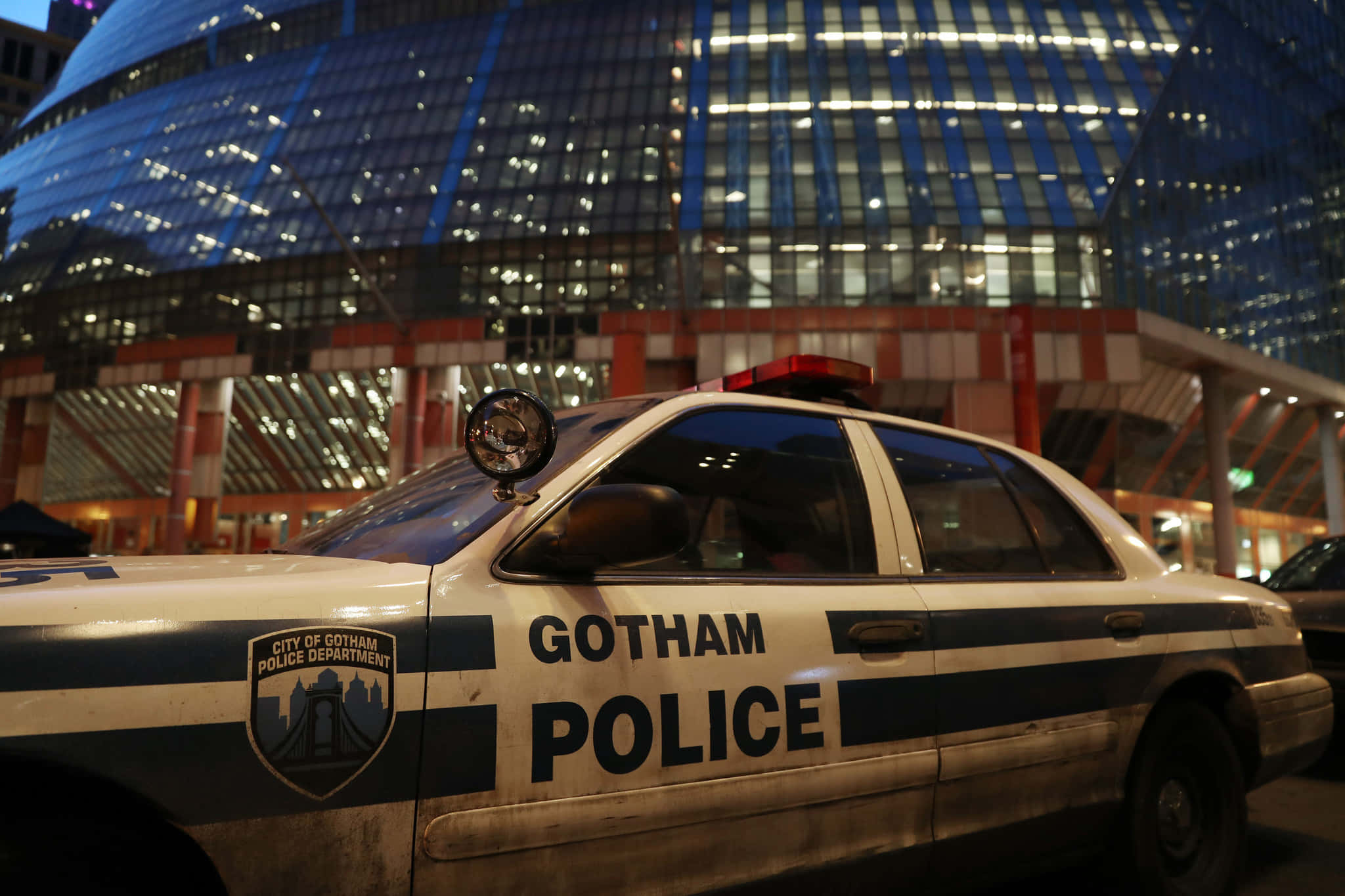 Gotham City Police Department 桌布 壁紙 — 一輛飽經風霜的高譚市警車在夜晚停泊在發光的未來主義摩天大樓前。