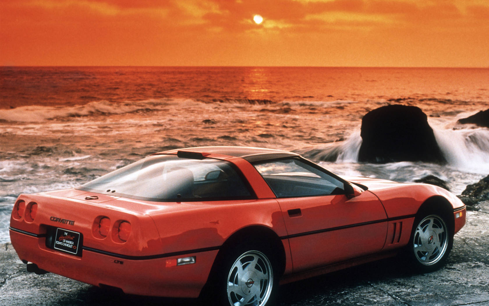 corvette 桌布 壁紙 — 一輛鮮橙色Corvette跑車停泊在岩石海岸上，海浪拍打岩石，背景是熾熱的日落天空。