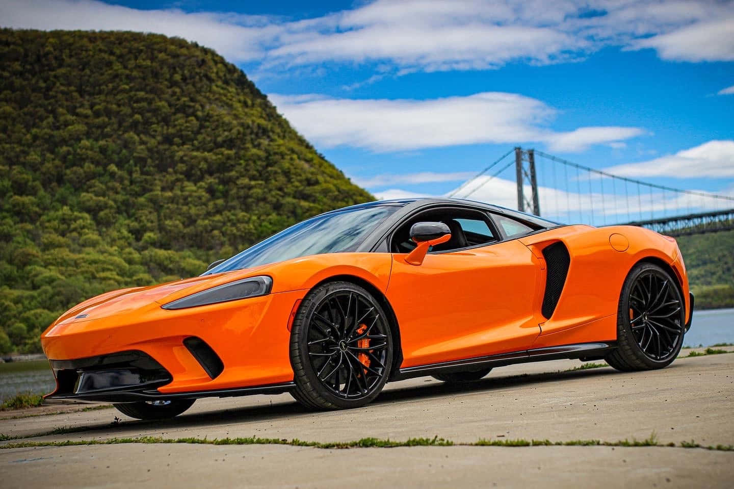 麥拉倫 桌布 壁紙 — 一輛鮮橙色McLaren跑車停泊在河邊的混凝土小徑上，背景是藍天下的一座綠色山丘和吊橋。