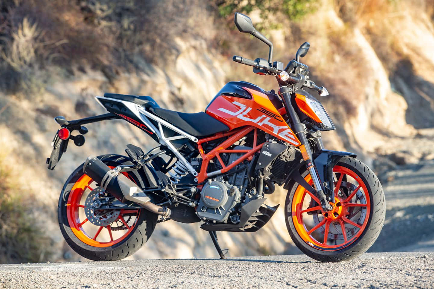  壁紙 — 一輛鮮橙色和黑色的KTM Duke摩托車停放在陽光明媚的碎石路上，背景是岩石山坡。