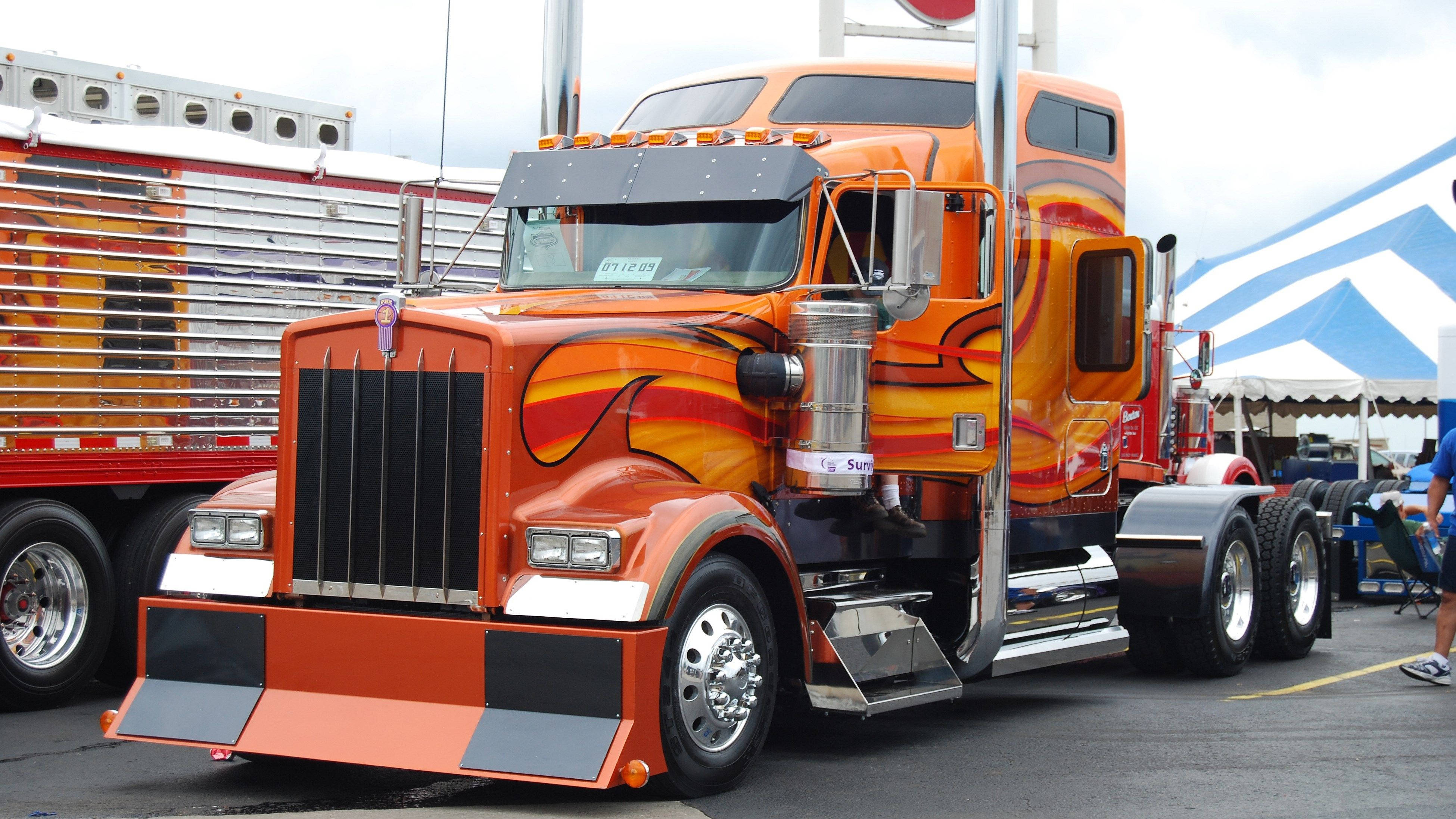 Kenworth 桌布 壁紙 — 一輛鮮橙色定制半掛卡車，帶有火焰貼花，停在藍白帳篷下的卡車展上。
