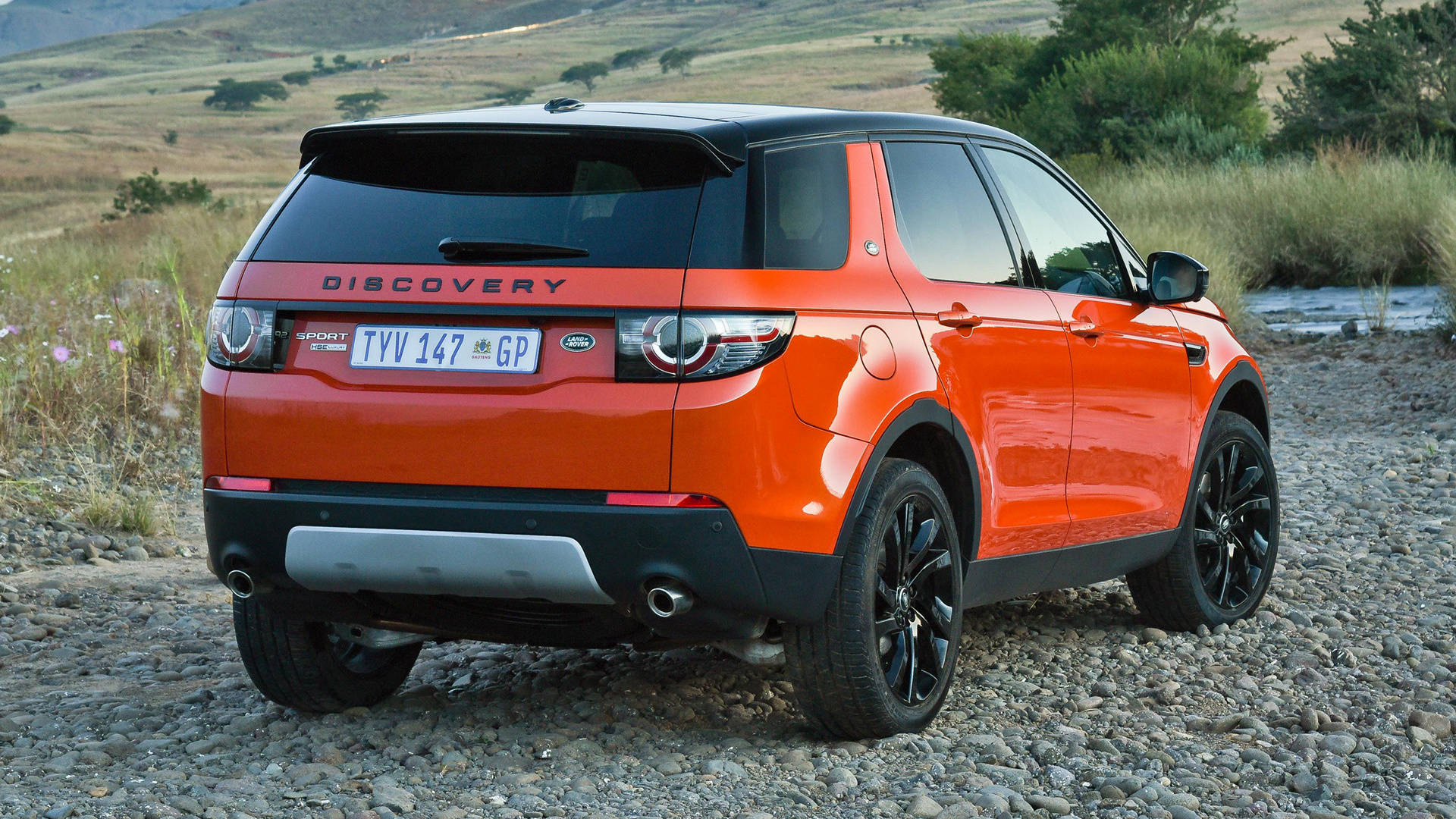land rover iPhone 桌布 壁紙 — 一輛鮮橙色的 Land Rover Discovery Sport 停泊在碎石路上，背景是起伏的山丘和乾草。