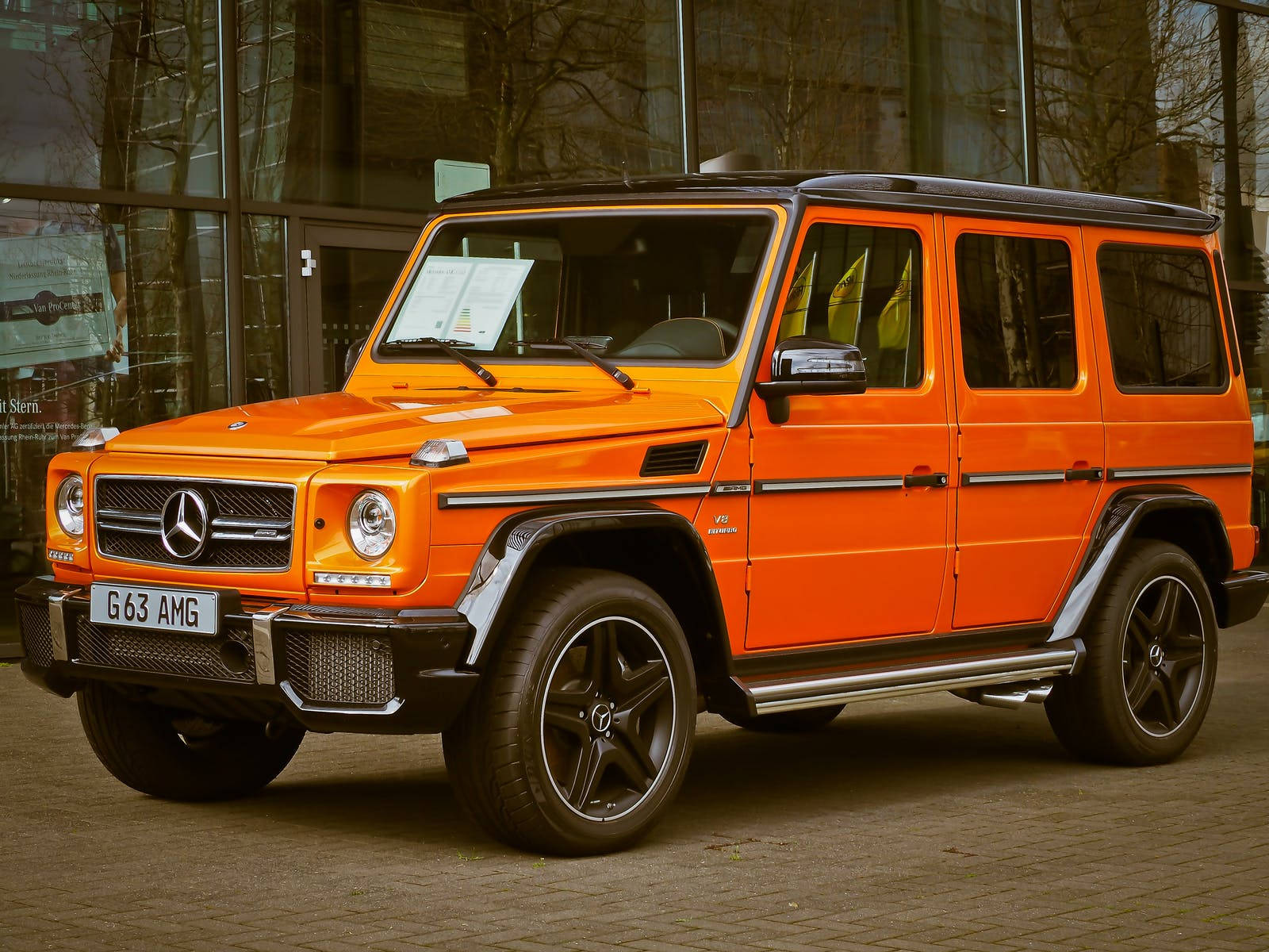 跑車 桌布 壁紙 — 一輛鮮橙色的 Mercedes G63 AMG SUV 停放在現代玻璃建築前的鋪砌地面上。